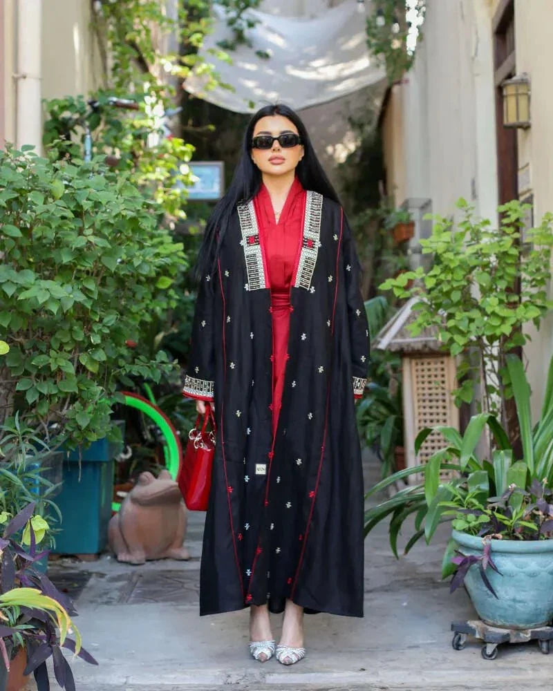 Woman wearing a black linen abaya with pearl embroidery and red piping over a crimson inner dress, accessorized with sunglasses and a red handbag, standing outdoors.