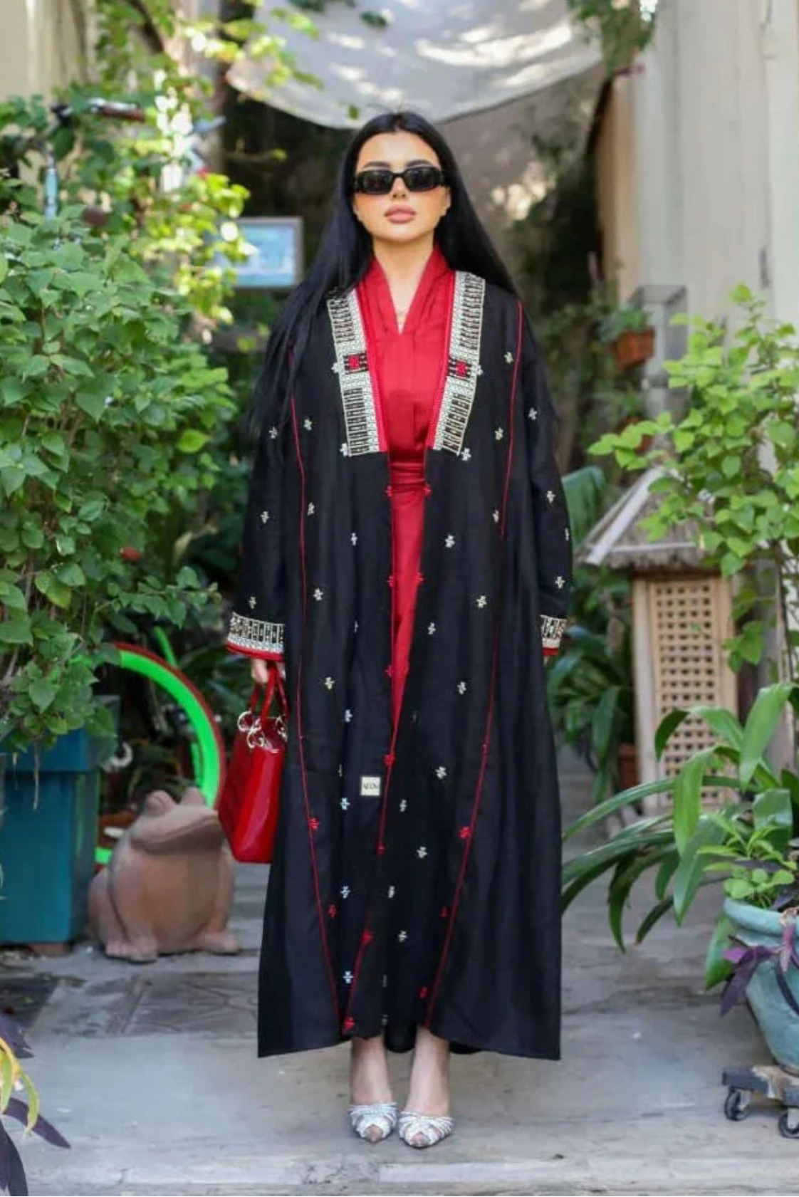 Woman wearing a black linen abaya with pearl embroidery and red piping over a crimson inner dress, accessorized with sunglasses and a red handbag, standing outdoors.