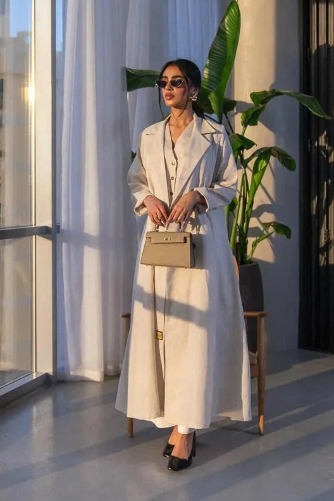 Woman wearing a beige linen blazer-style abaya with classic lapel collar and front buttons in a modern, sunlit room