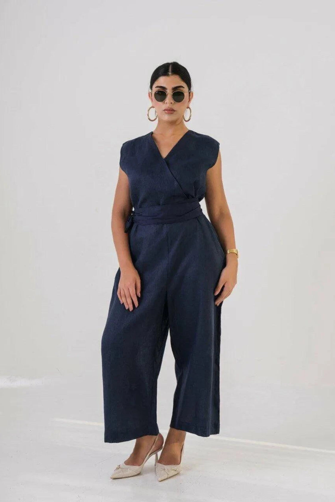 Woman wearing navy linen jumpsuit with V-neckline, fabric belt, and wide-leg pants, styled with sunglasses and heels