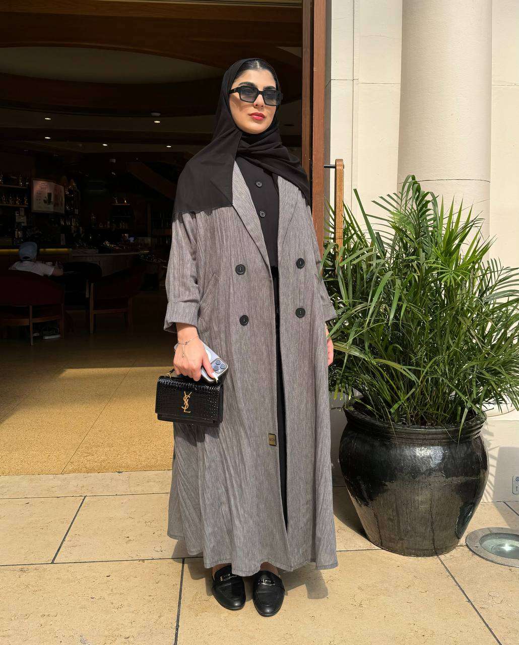 Woman wearing a grey modern abaya with double-breasted buttons and black hijab standing outdoors next to a plant pot