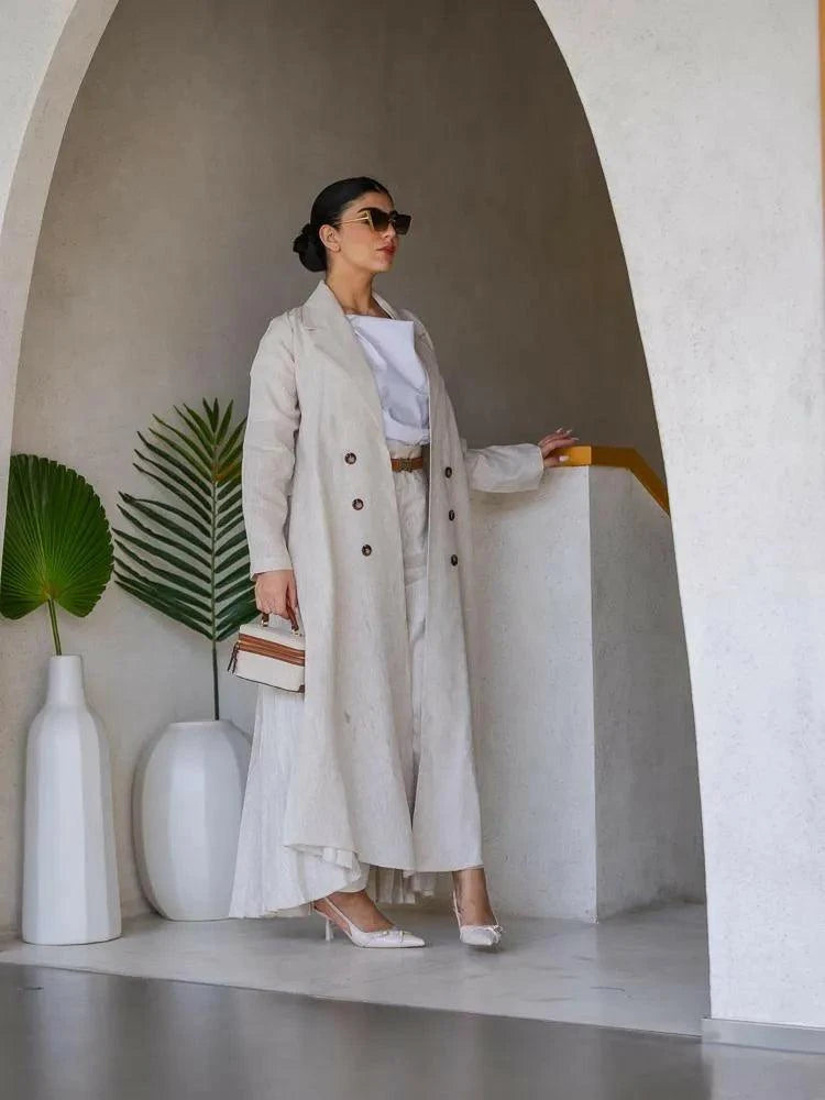 Woman wearing Whisper Beige linen abaya with pleated chiffon panels and double-breasted buttons, styled with a white top, beige skirt, and heels.