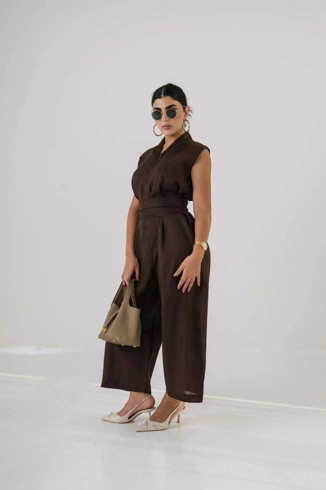 Woman wearing dark brown linen jumpsuit with sleeveless design, holding beige handbag and wearing sunglasses and heels