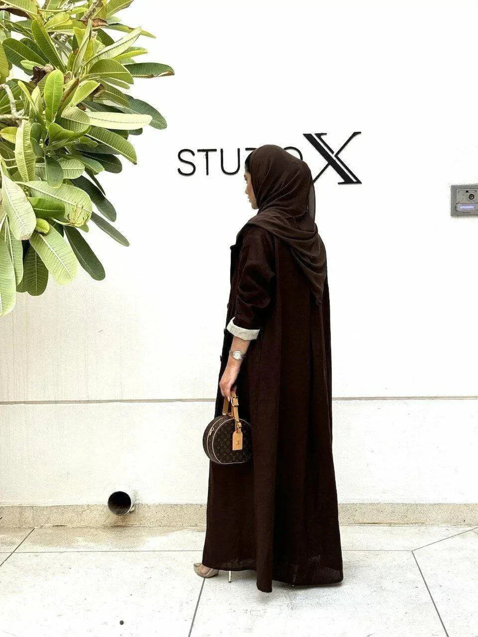 woman wearing a dark brown tailored linen blazer styled as a long coat with a matching headscarf and white outfit underneath standing against a white wall with a green plant nearby - dark brown blazer