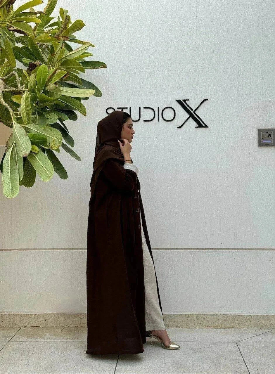 woman wearing a dark brown tailored linen blazer styled as a long coat with a matching headscarf and white outfit underneath standing against a white wall with a green plant nearby - dark brown blazer