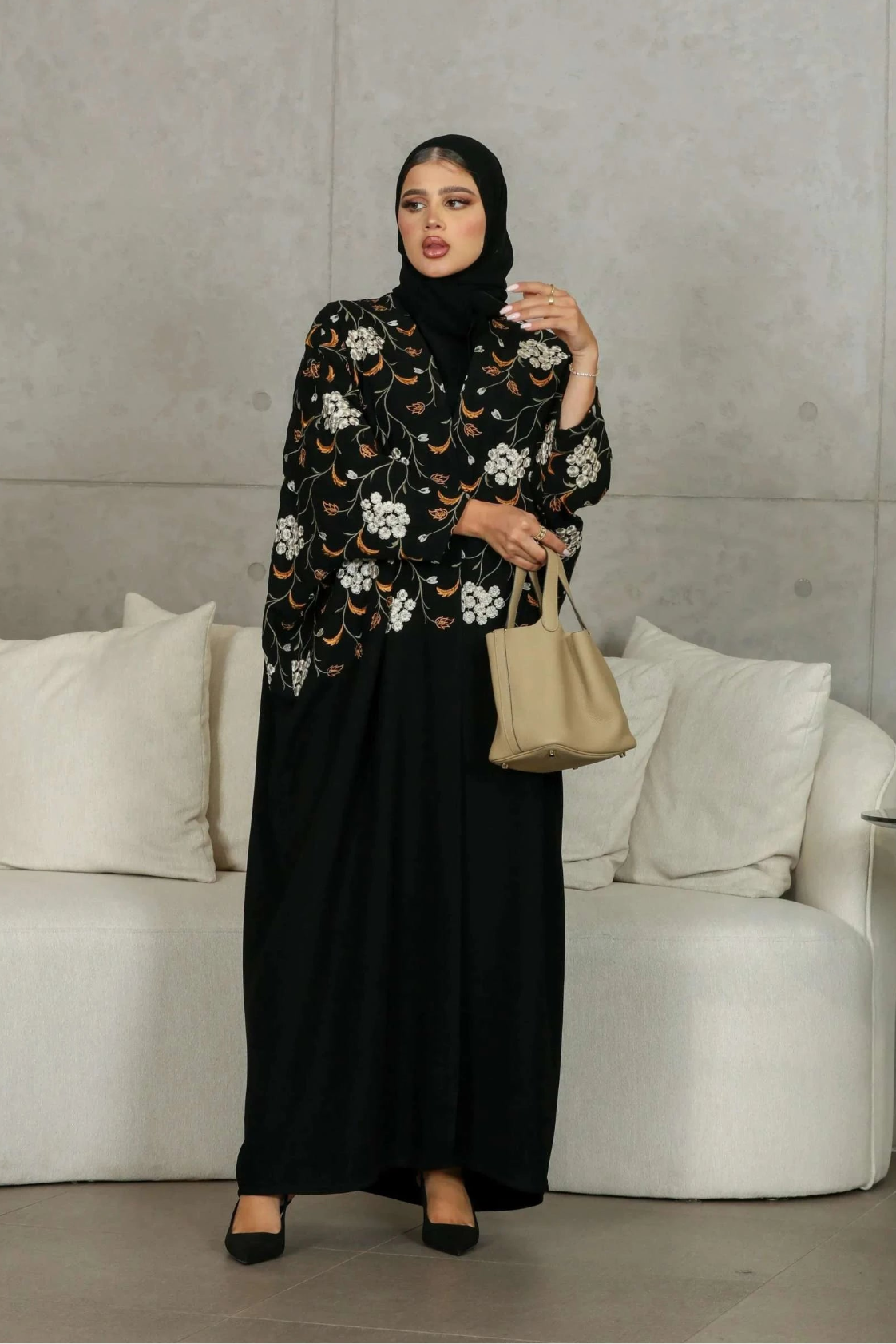 Elegant black abaya with white and earthy orange floral embroidery on bodice and sleeves, worn with a beige handbag in a modern setting