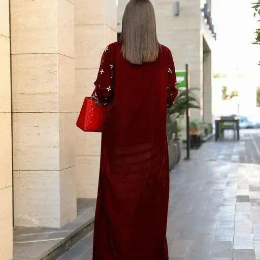 Back view of a luxurious maroon velvet abaya with delicate embroidery on sleeves worn by a woman carrying a red handbag outdoors.
