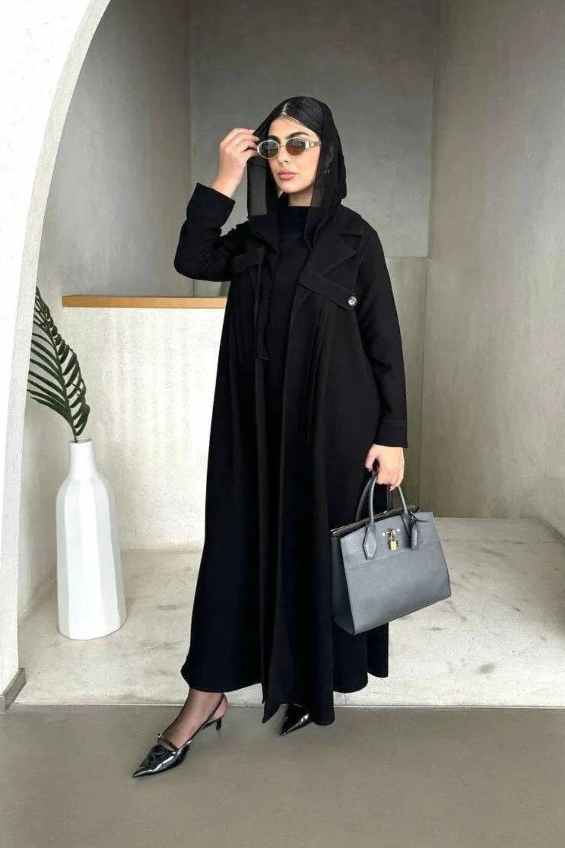 Woman wearing Obscia black abaya with blazer-style trench collar and structured shoulder flaps, holding a gray handbag and sunglasses.