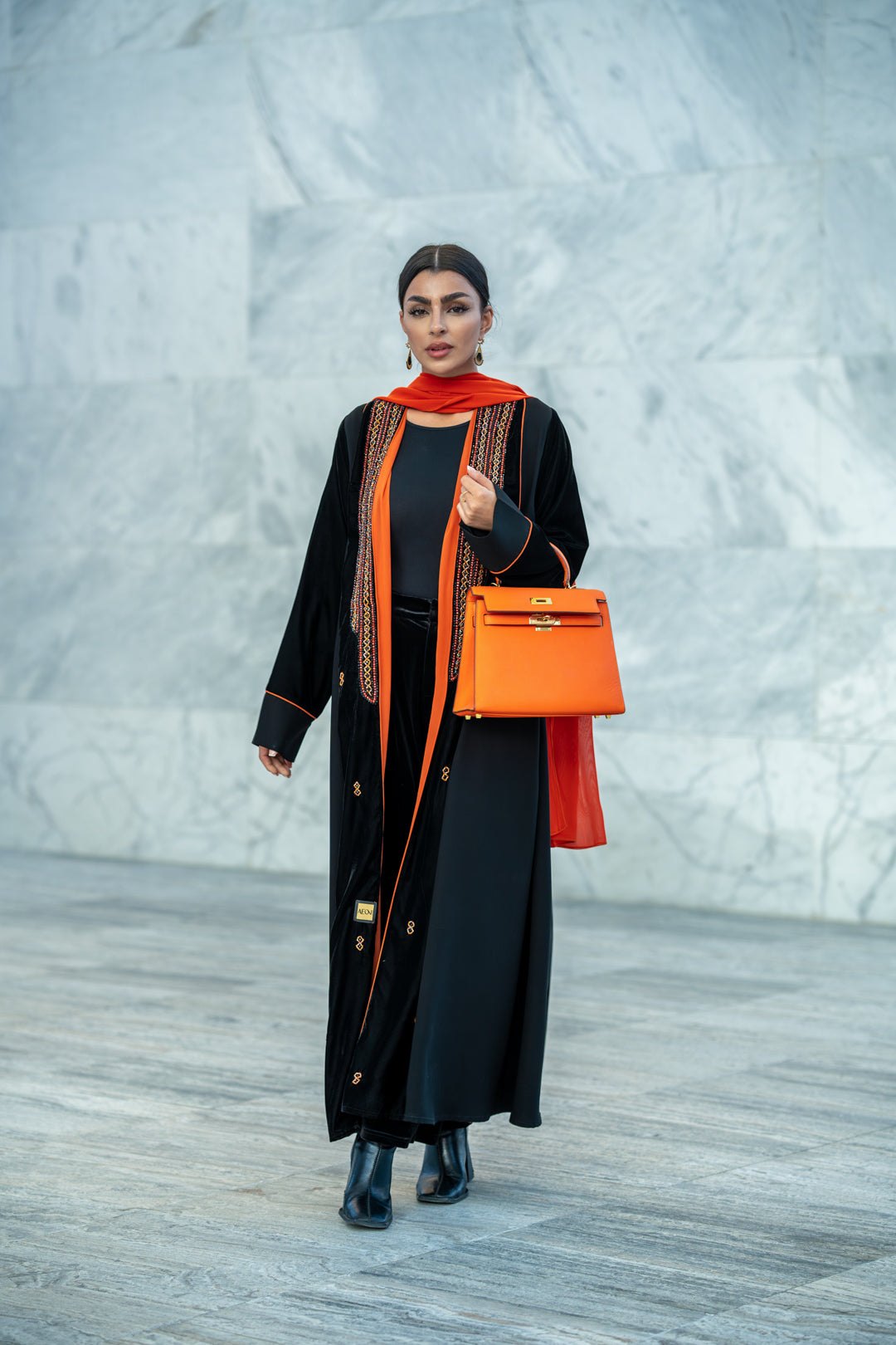 Black velvet abaya with vibrant orange accents, hand embroidery, and contrast piping worn with matching orange scarf and handbag.