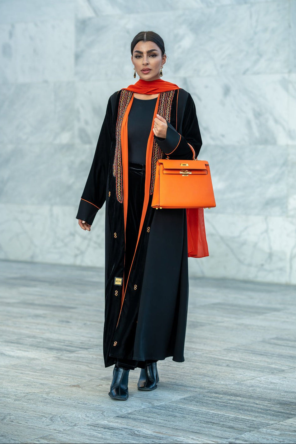 Black velvet abaya with vibrant orange accents, hand embroidery, and contrast piping worn with matching orange scarf and handbag.