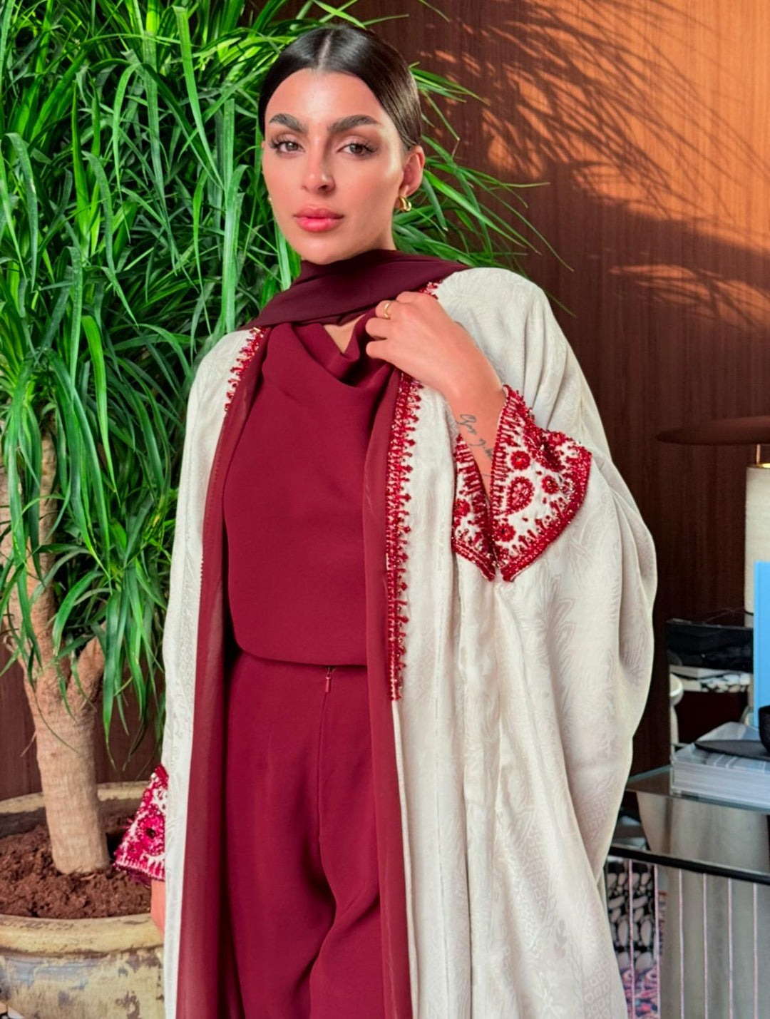 Woman wearing Harir Kubra Ivory Abaya with burgundy embroidery on sleeves and front edges in elegant indoor setting