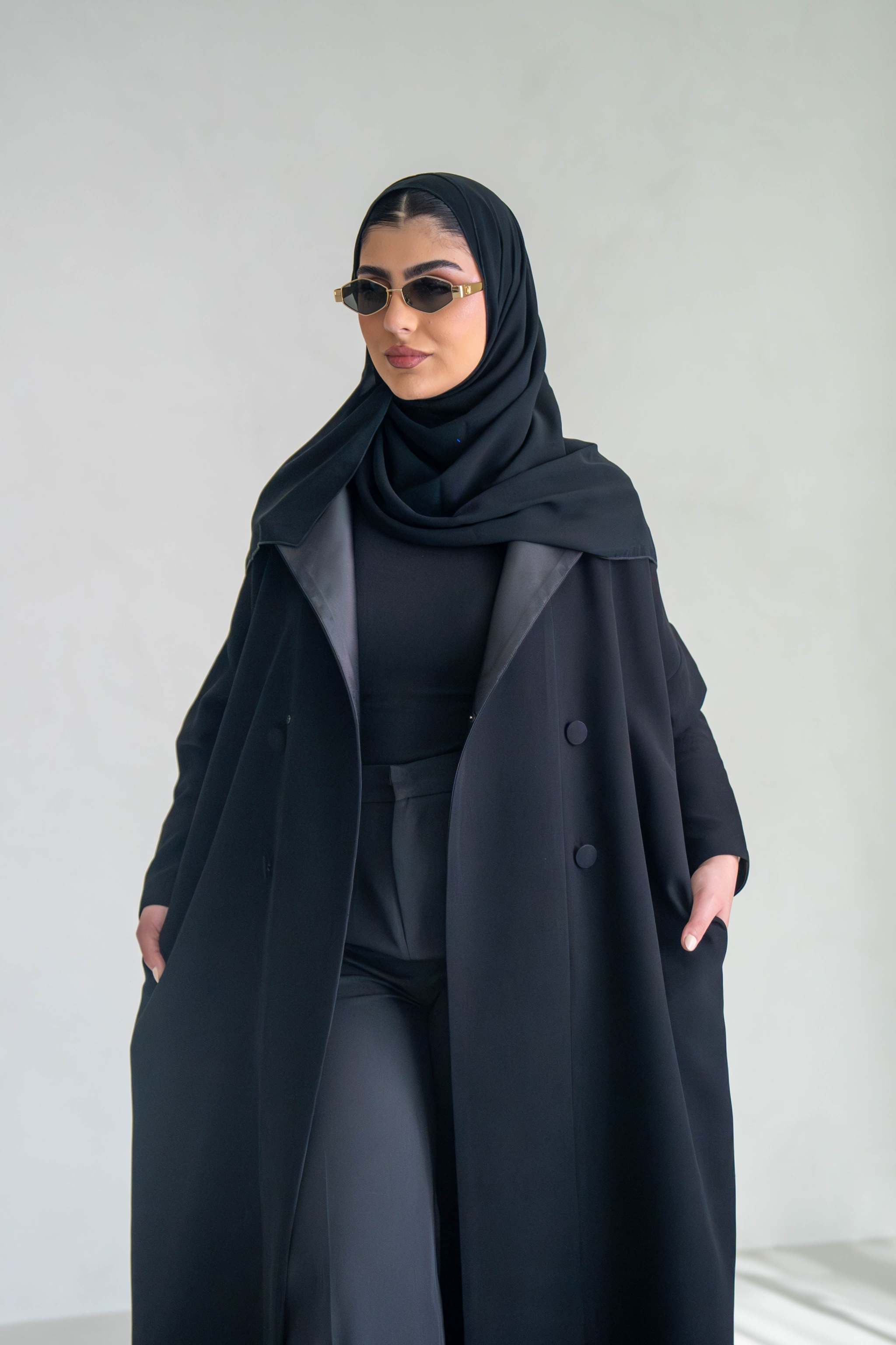 Woman wearing thick black crepe abaya with sunglasses and hijab against a plain background