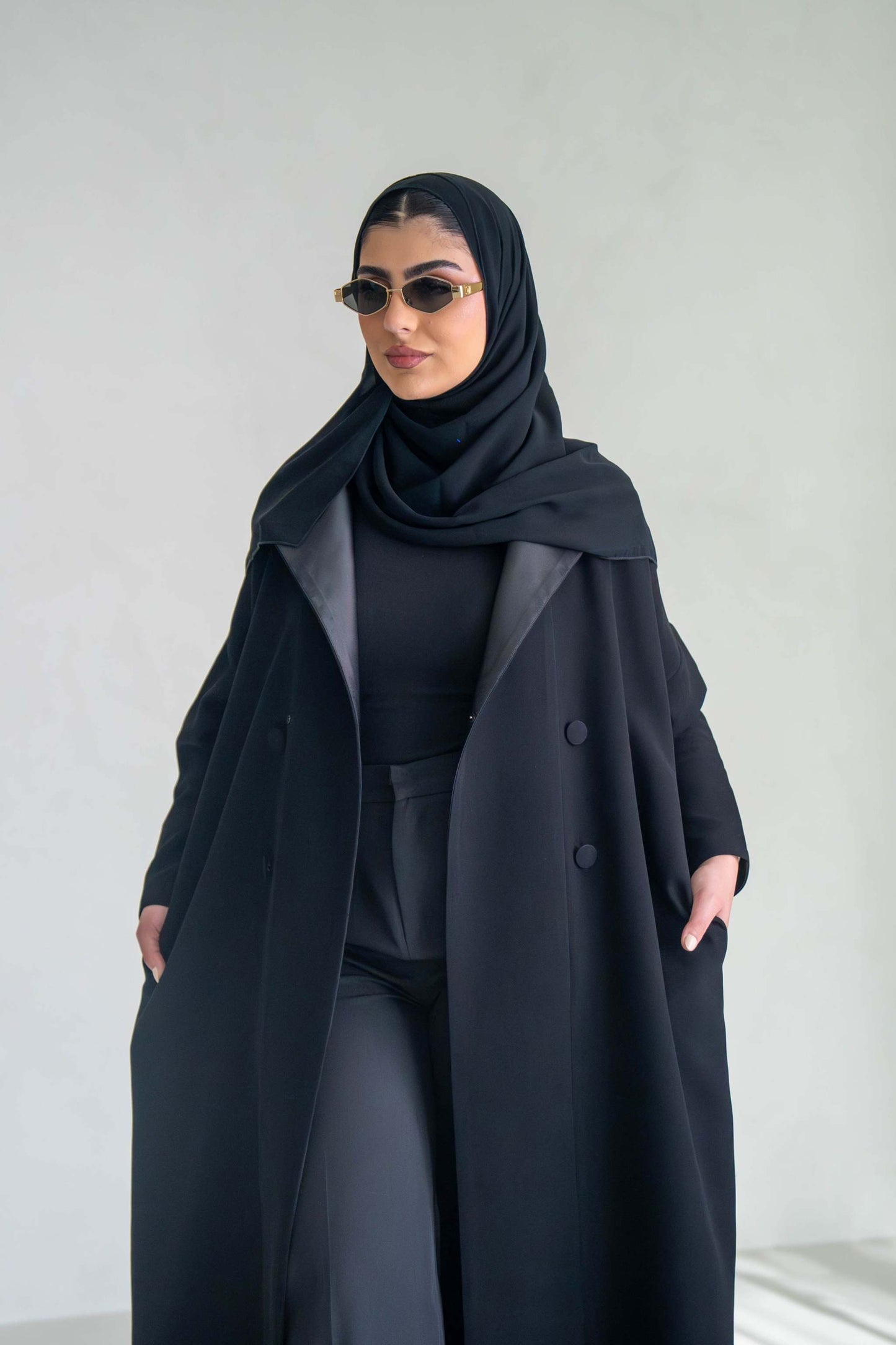 Woman wearing thick black crepe abaya with sunglasses and hijab against a plain background