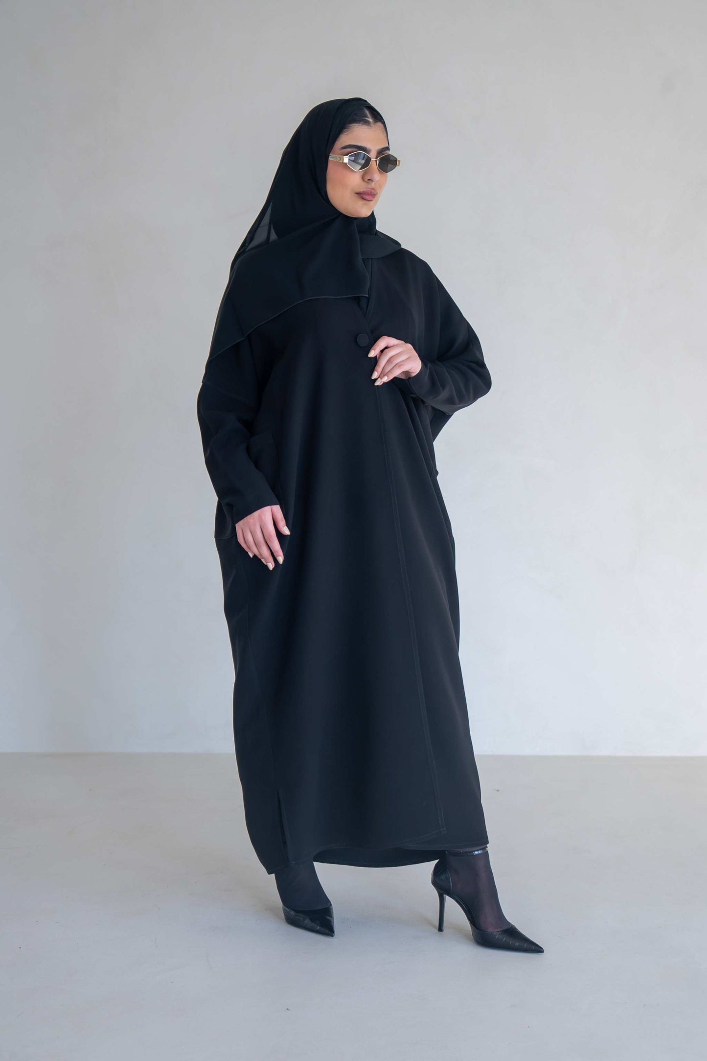 Woman wearing a classic black abaya with wide sleeves and side pockets, styled with black heels and sunglasses.