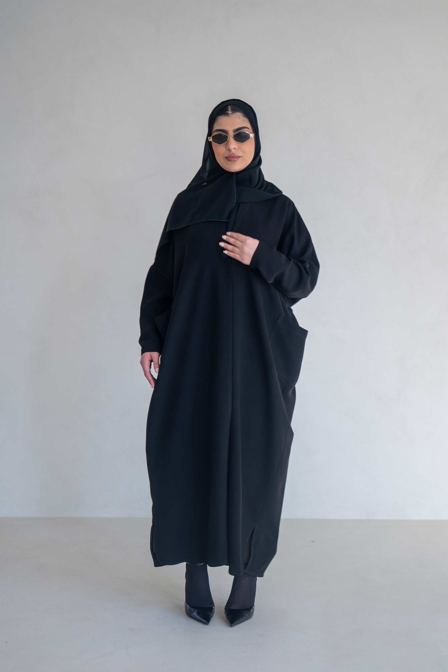 woman wearing classic black abaya with wide sleeves and side pockets standing against plain background