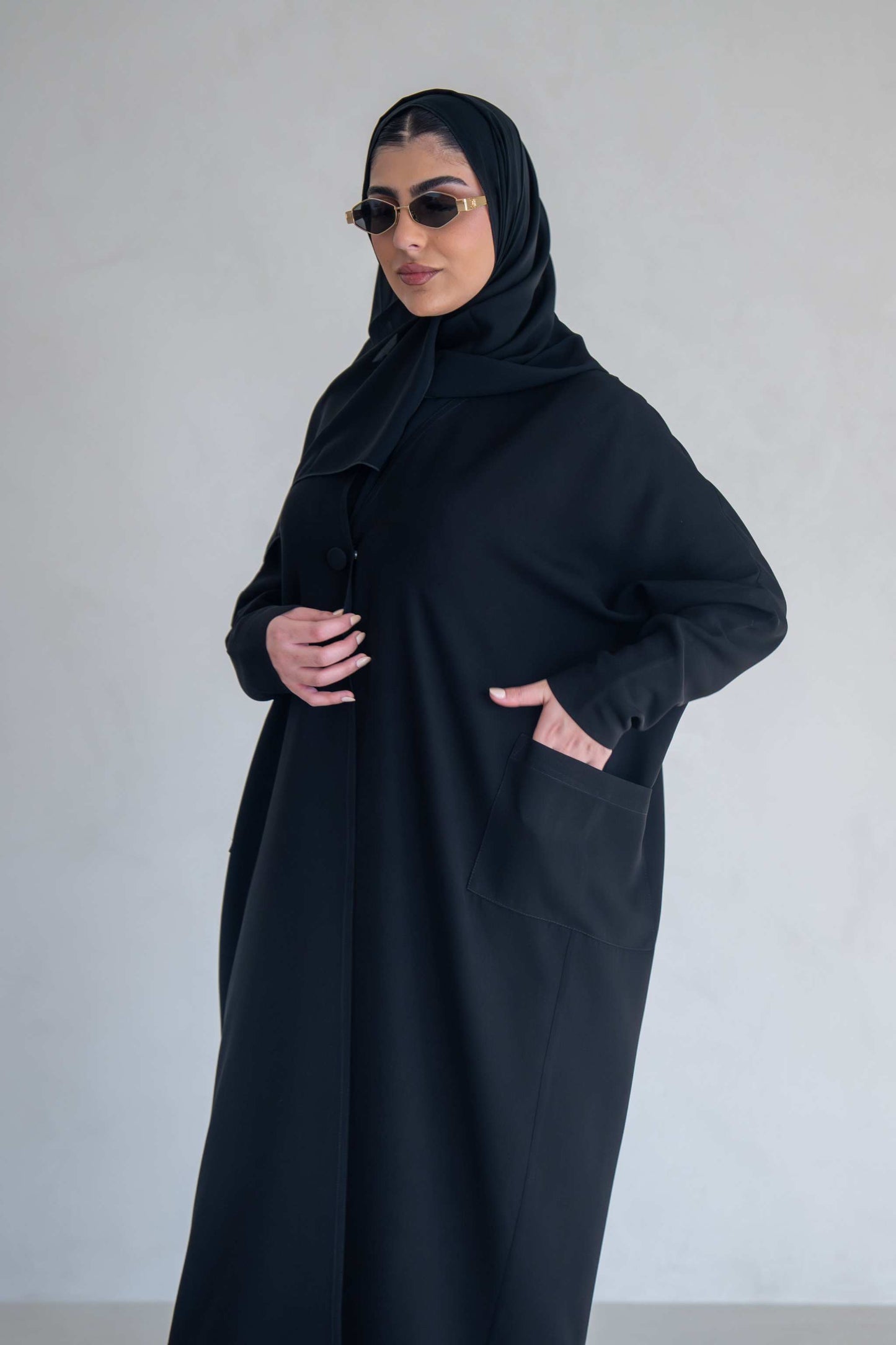 woman wearing classic black abaya with wide sleeves and side pockets standing against plain background