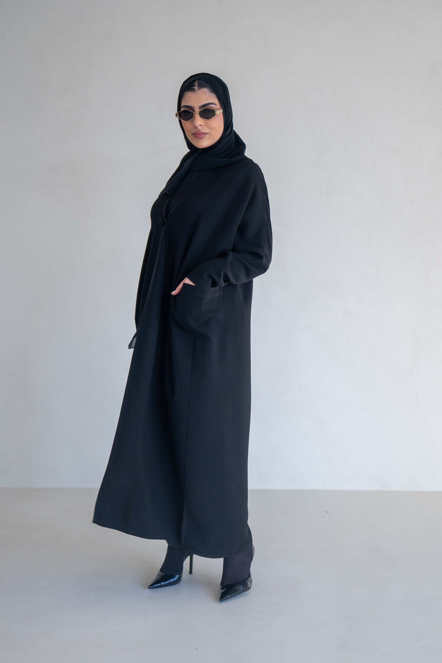 woman wearing classic black abaya with wide sleeves and side pockets standing against plain background