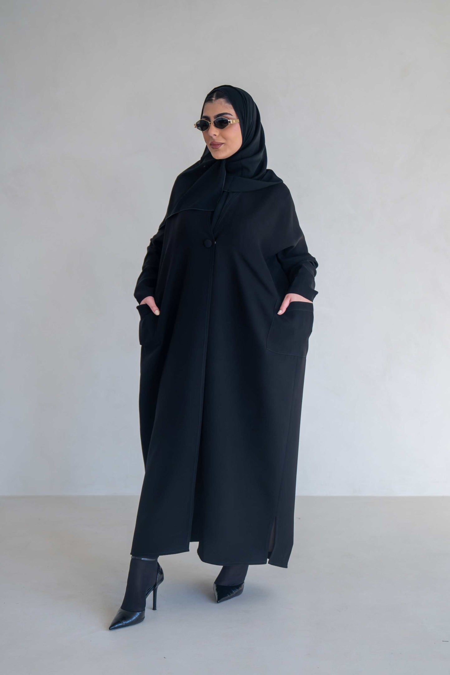 woman wearing classic black abaya with wide sleeves and side pockets standing against plain background