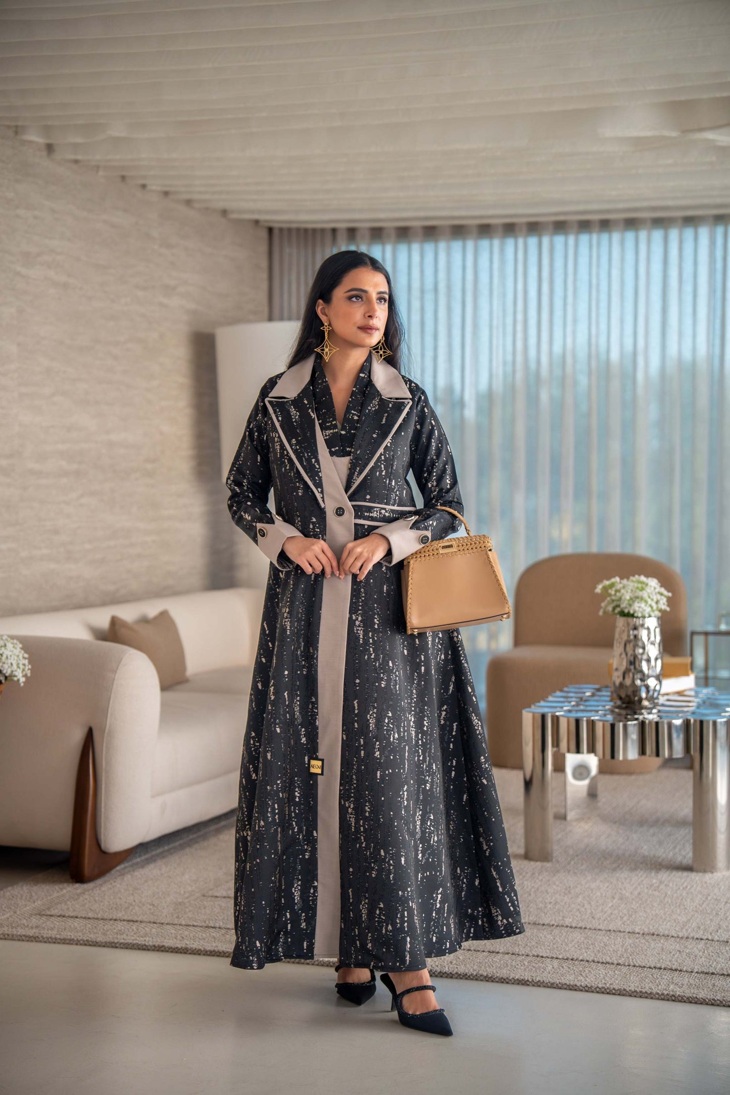 woman wearing elegant black and beige patterned Ashira abaya holding tan handbag in modern living room