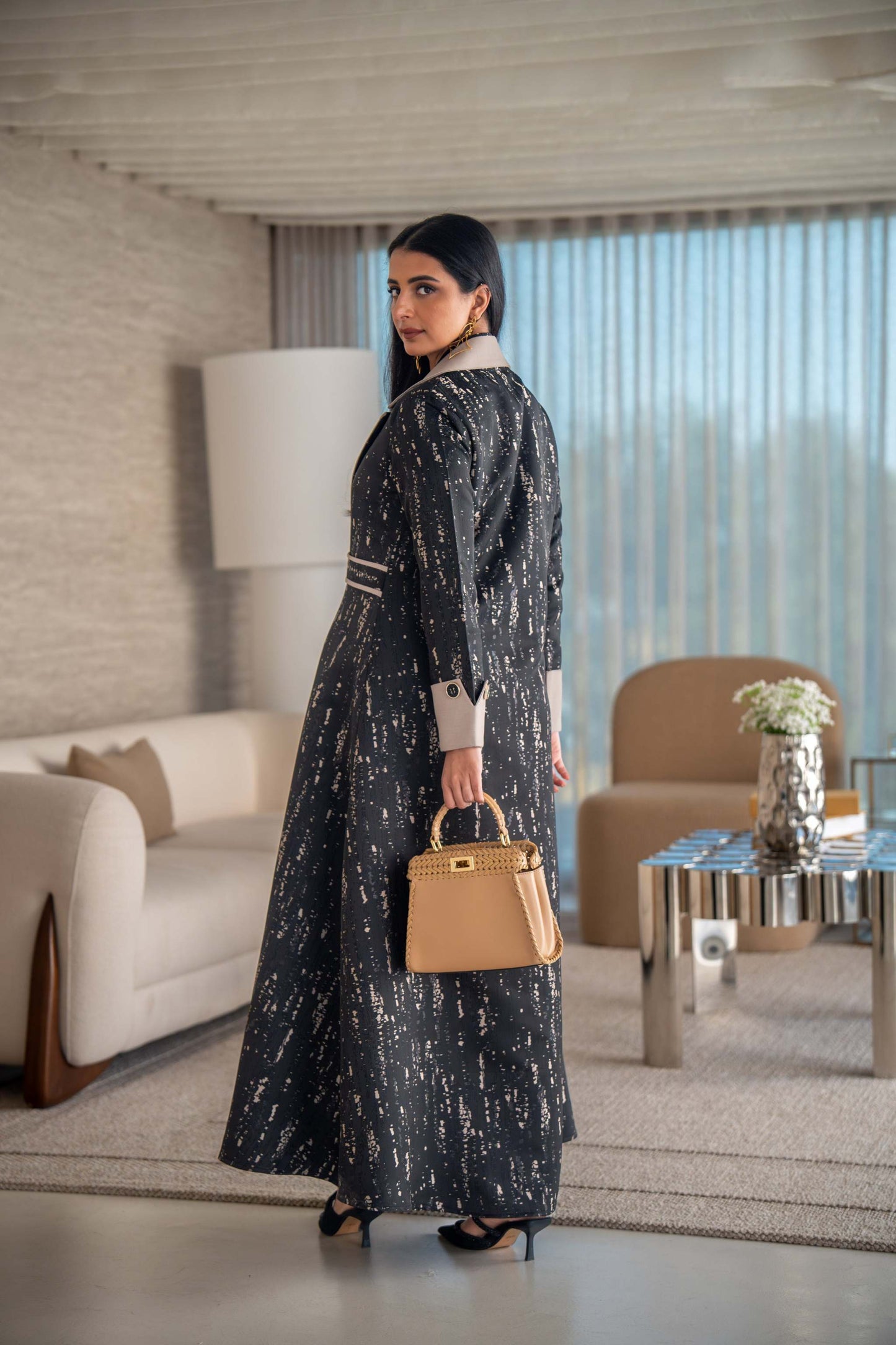 woman wearing elegant black and beige patterned Ashira abaya holding tan handbag in modern living room