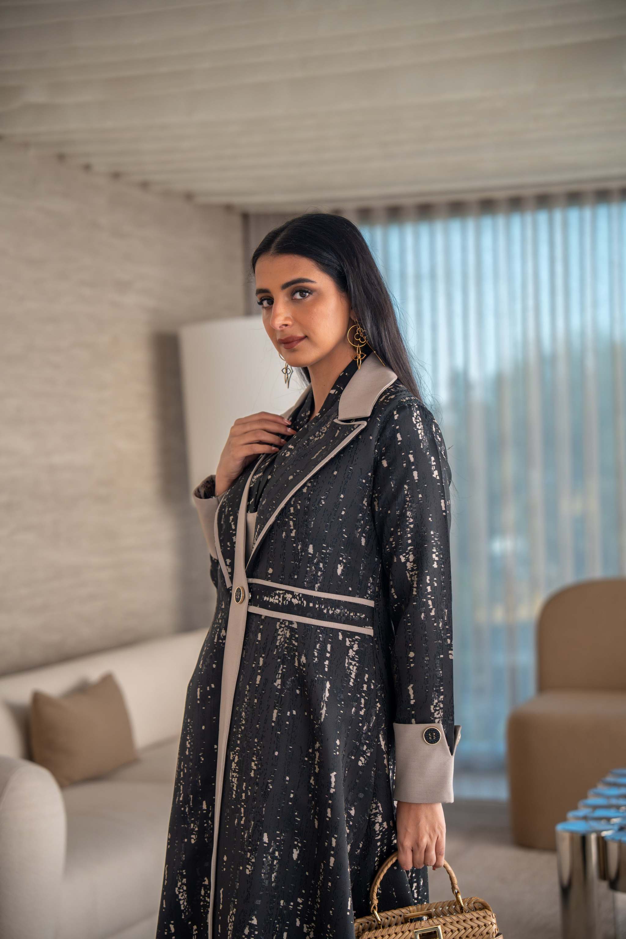 woman wearing elegant black and beige patterned Ashira abaya holding tan handbag in modern living room