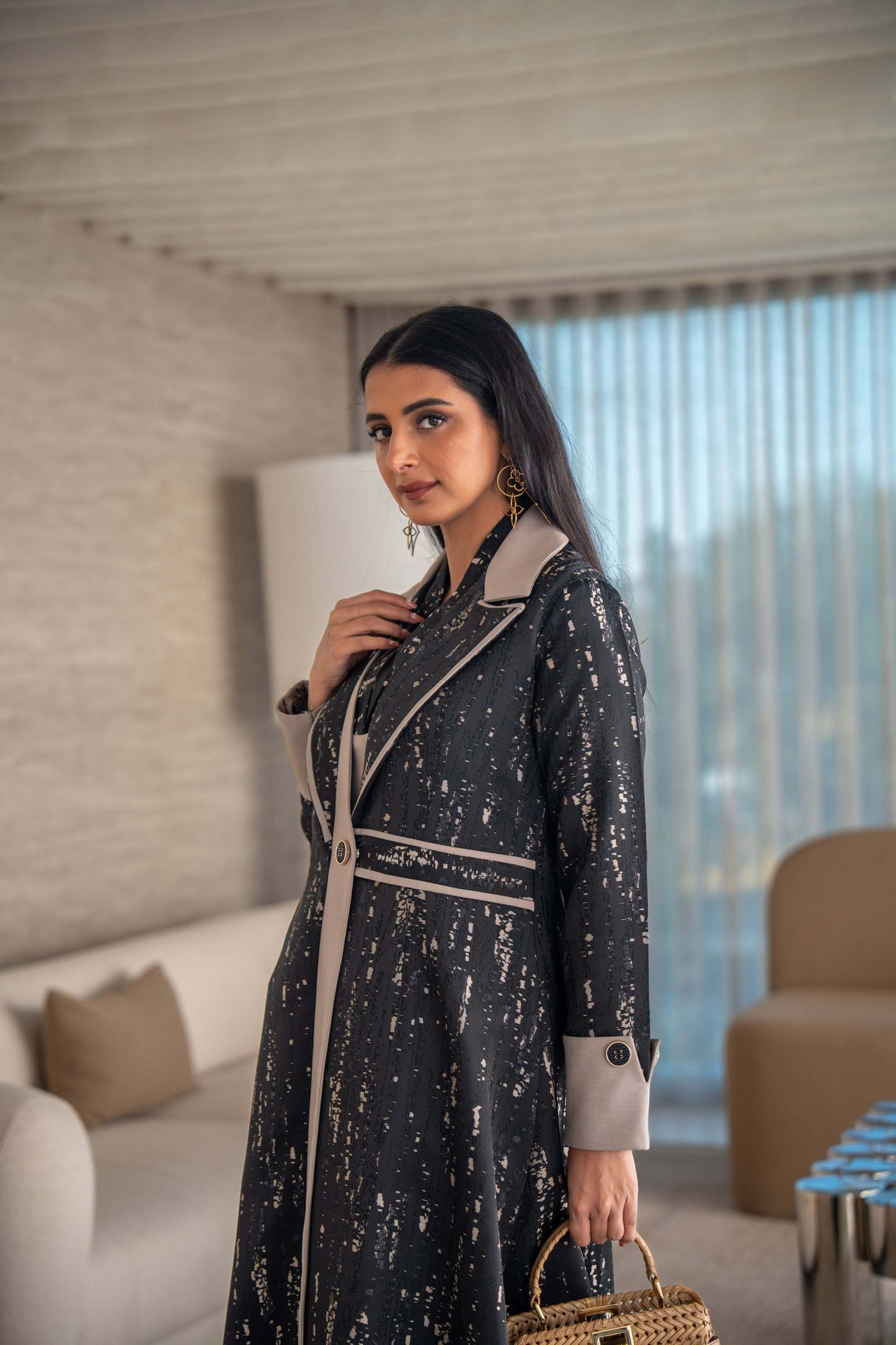 woman wearing elegant black and beige patterned Ashira abaya holding tan handbag in modern living room