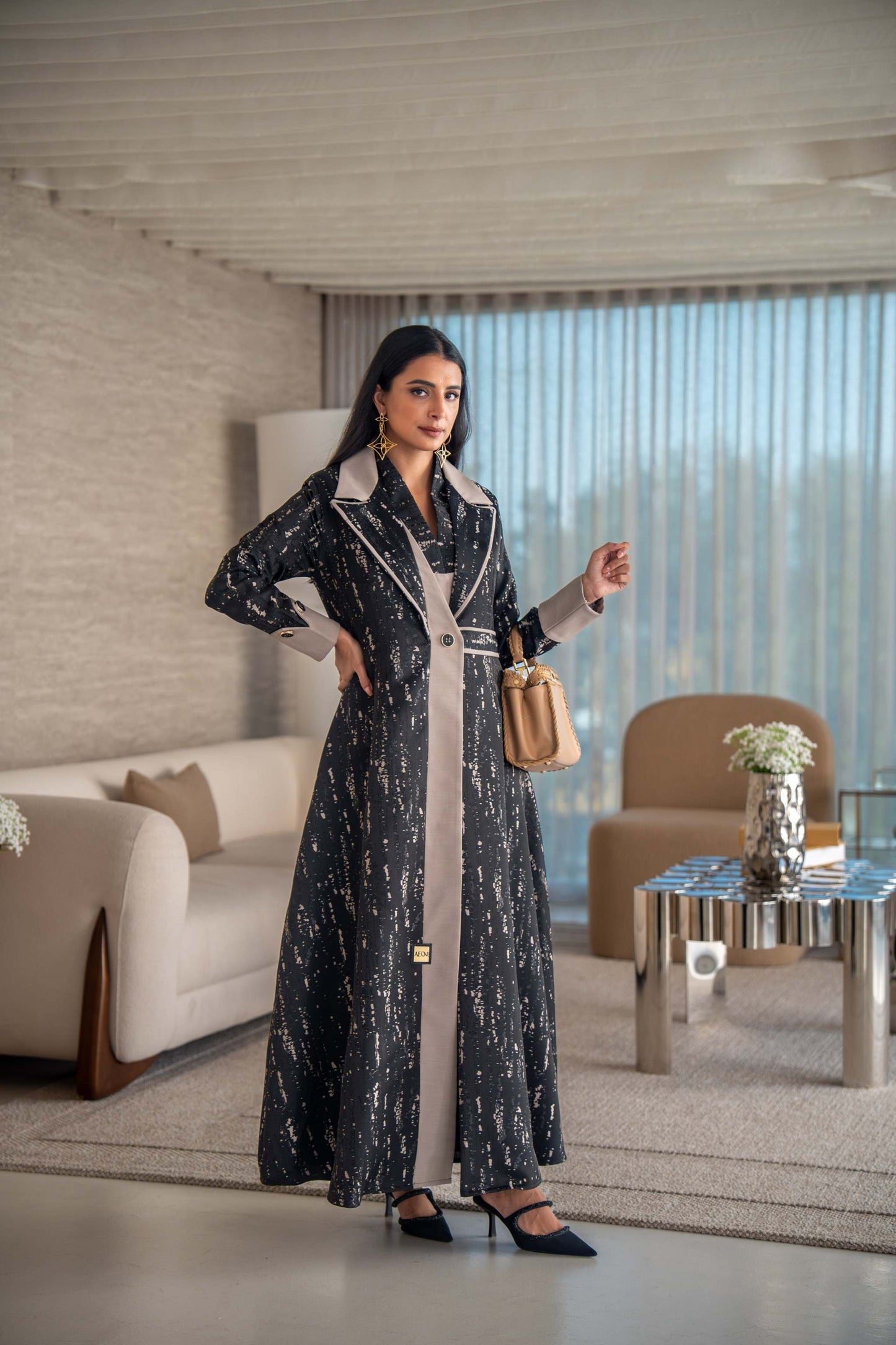 woman wearing elegant black and beige patterned Ashira abaya holding tan handbag in modern living room