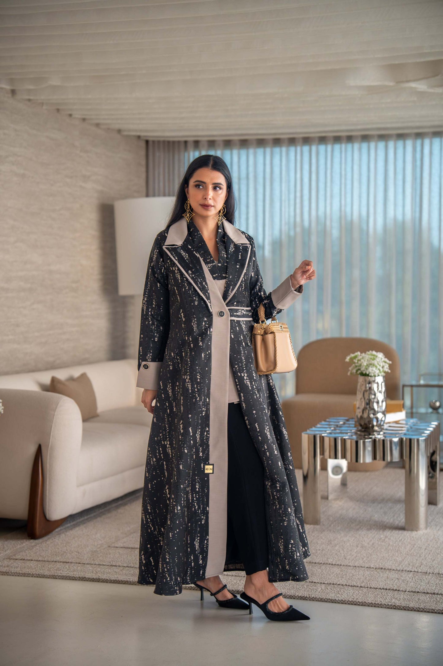 woman wearing elegant black and beige patterned Ashira abaya holding tan handbag in modern living room