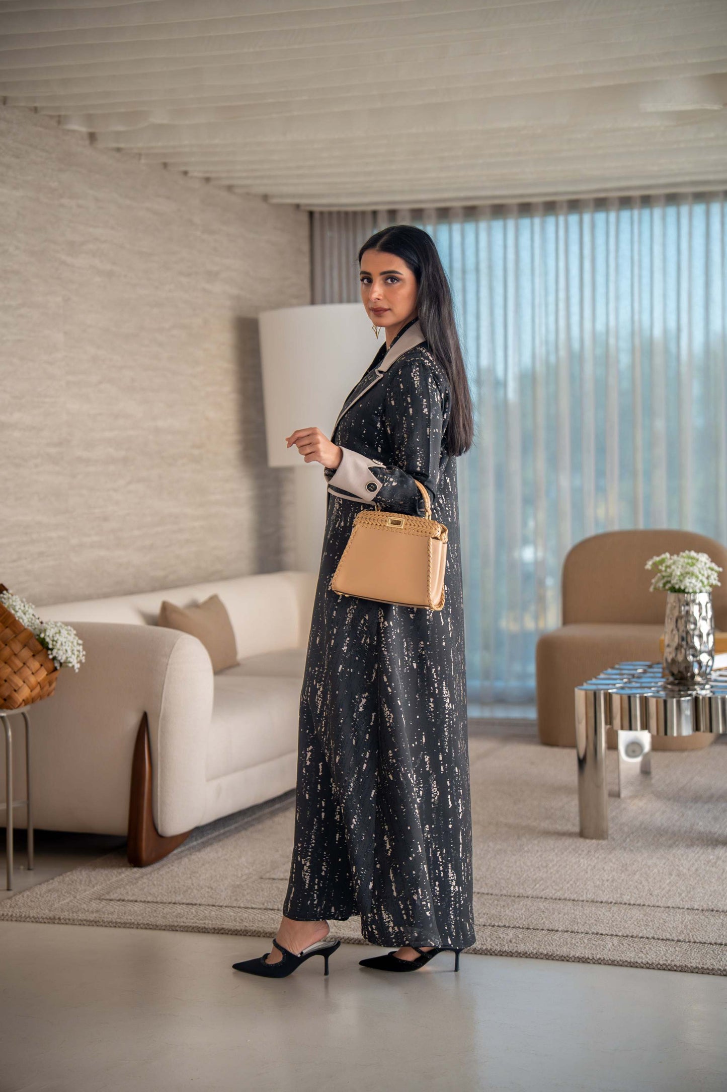 woman wearing elegant black and beige patterned Ashira abaya holding tan handbag in modern living room