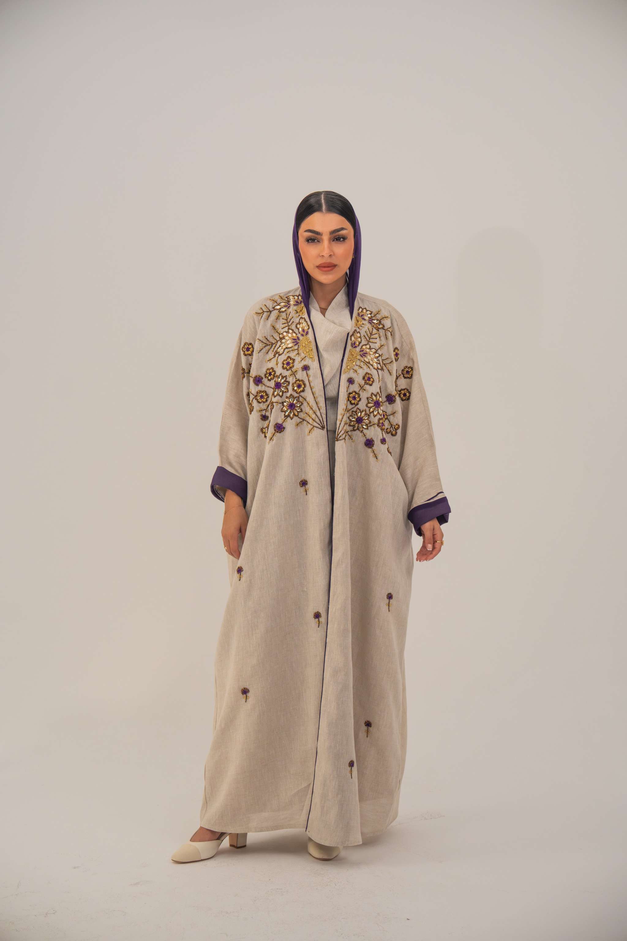 Woman wearing a beige open-front bisht abaya with gold and purple floral beadwork, purple chiffon lining, and relaxed fit.