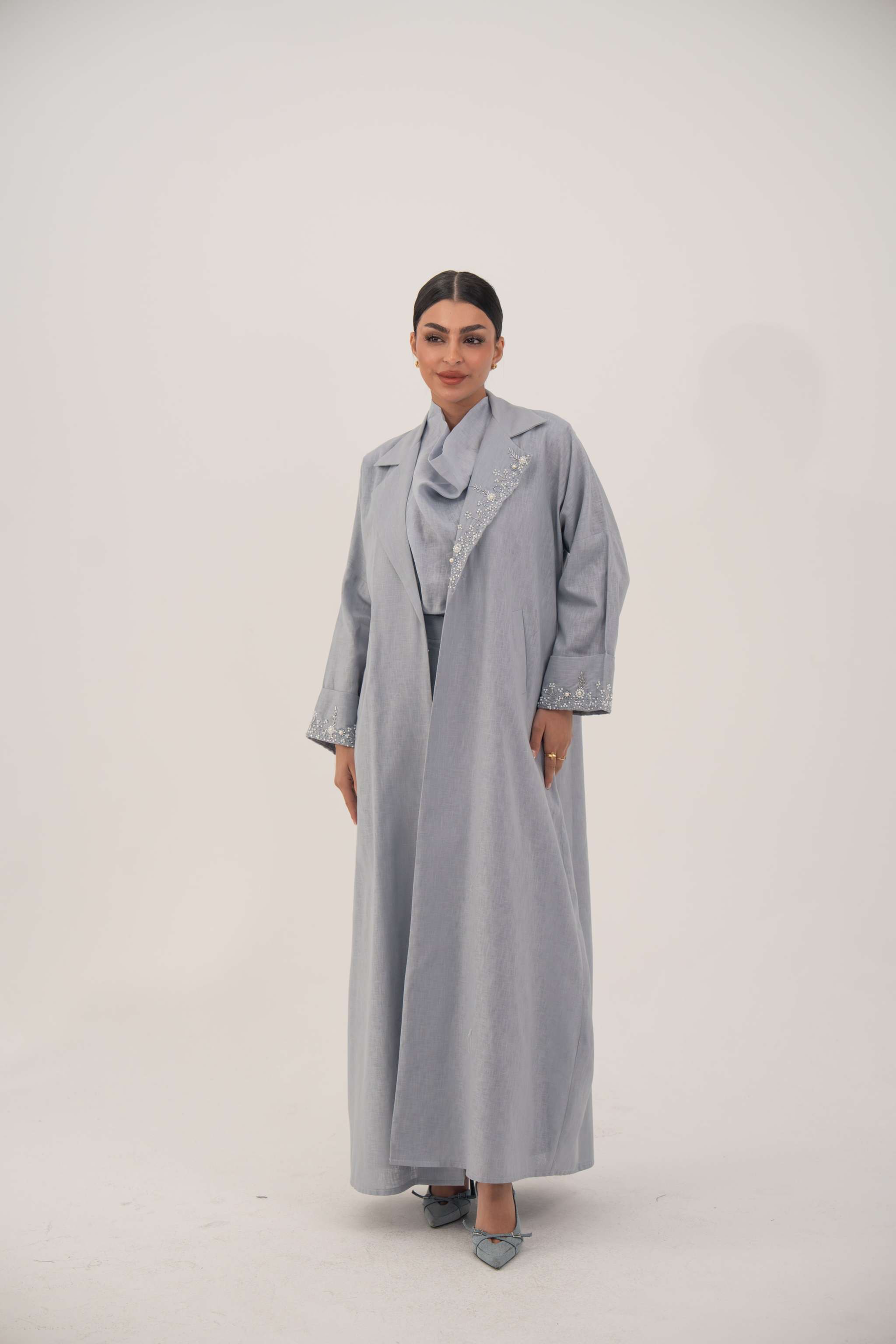Woman wearing a lightweight soft blueish gray modern blazer-style abaya with delicate white bead and crystal embellishments on the wide lapel and cuffs.