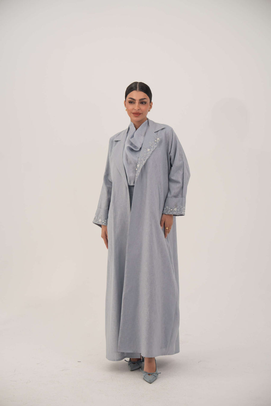 Woman wearing a lightweight soft blueish gray modern blazer-style abaya with delicate white bead and crystal embellishments on the wide lapel and cuffs.