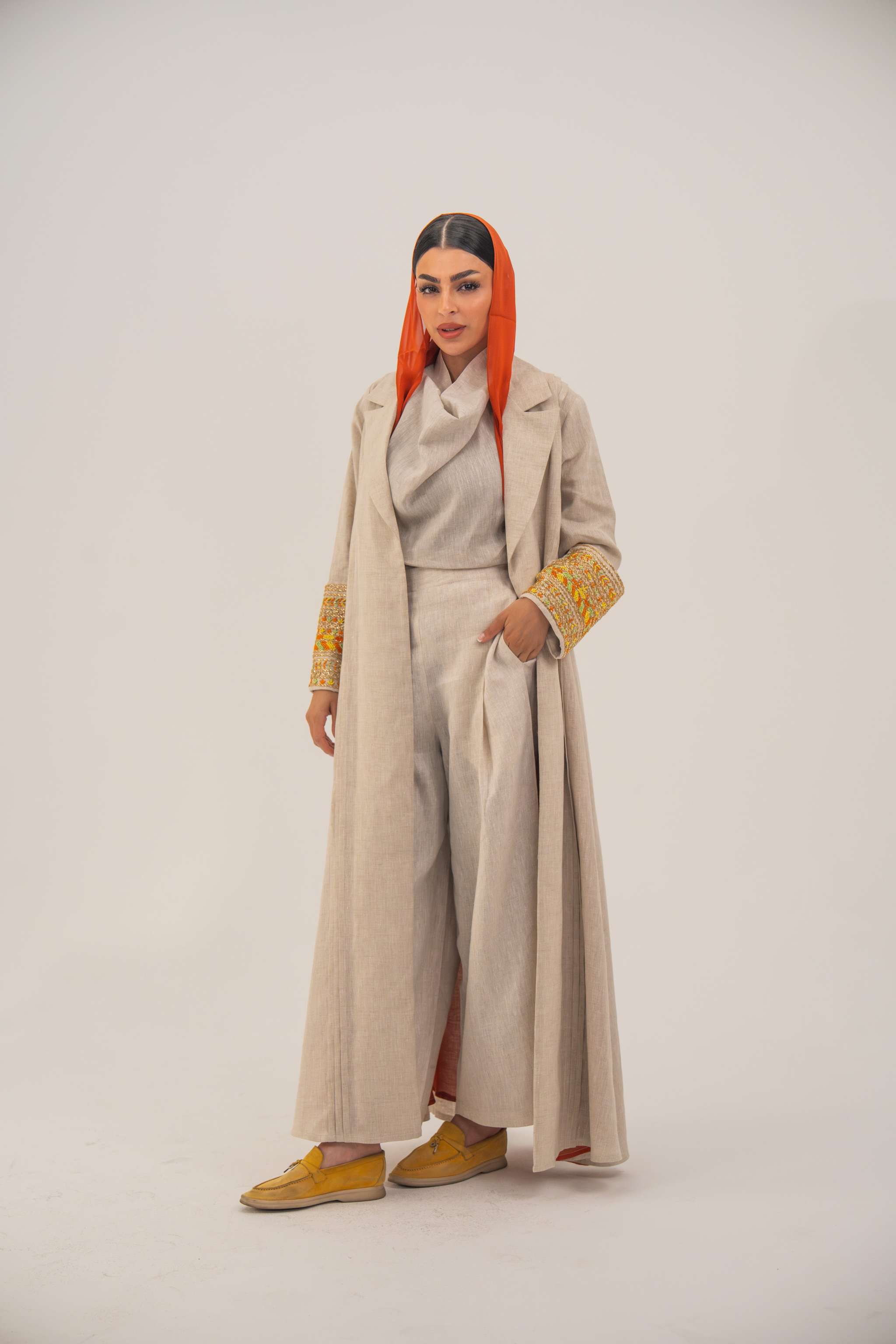 Woman wearing a breathable beige linen sophisticated abaya with tailored blazer-style cut and vibrant orange, gold, and green shak embroidery on the sleeves.