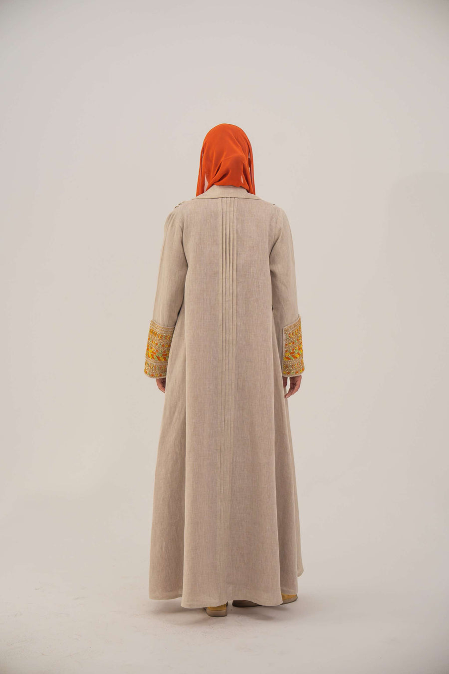 Back view of a beige linen blend abaya with wide lapels, orange, gold, and green embroidery on sleeves, and an orange chiffon inner lining, worn with an orange headscarf.-sophisticated abaya