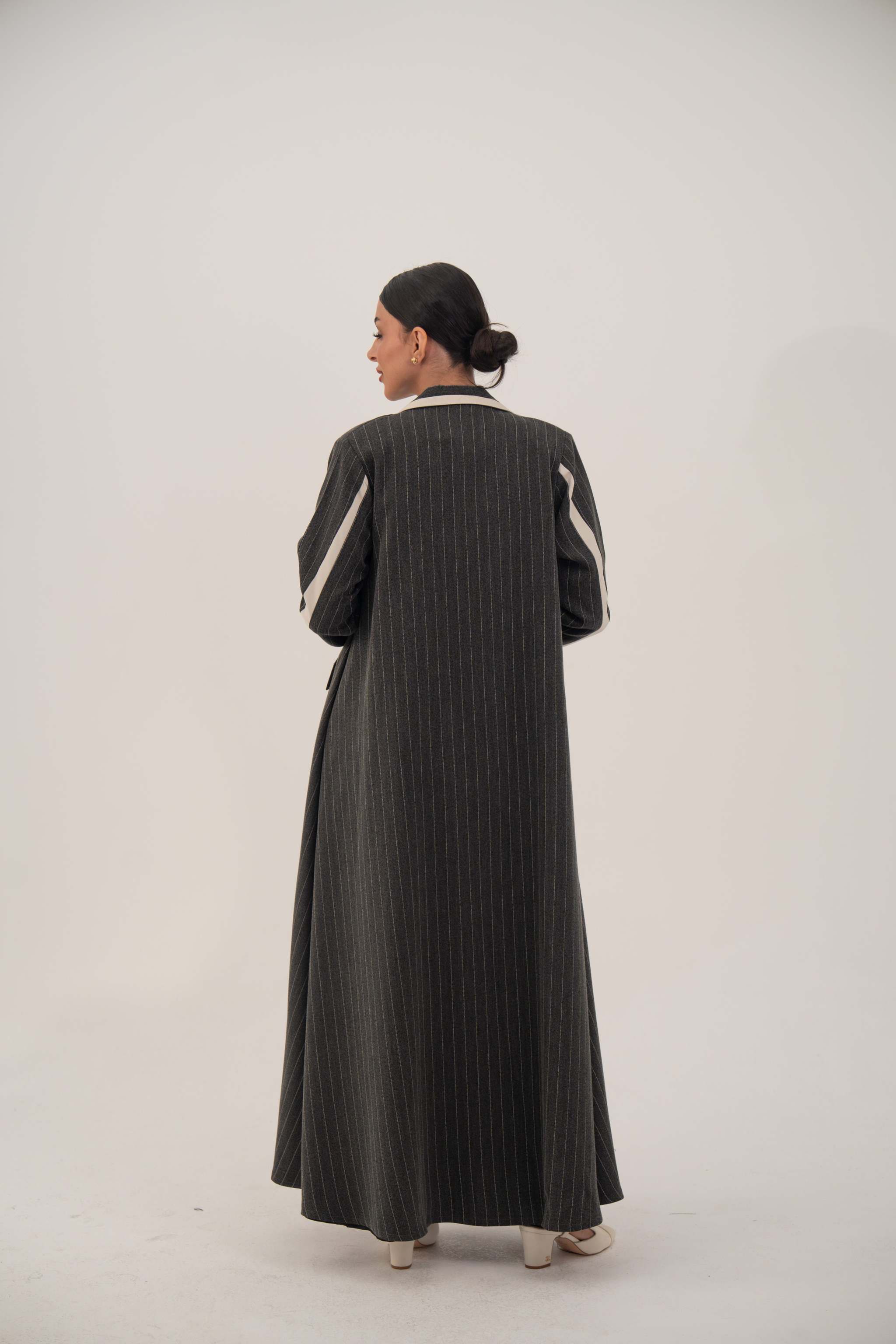 Back view of a woman wearing a dark pinstriped blazer-style abaya with off-white inner cuffs and shoulder details, standing against a plain background.