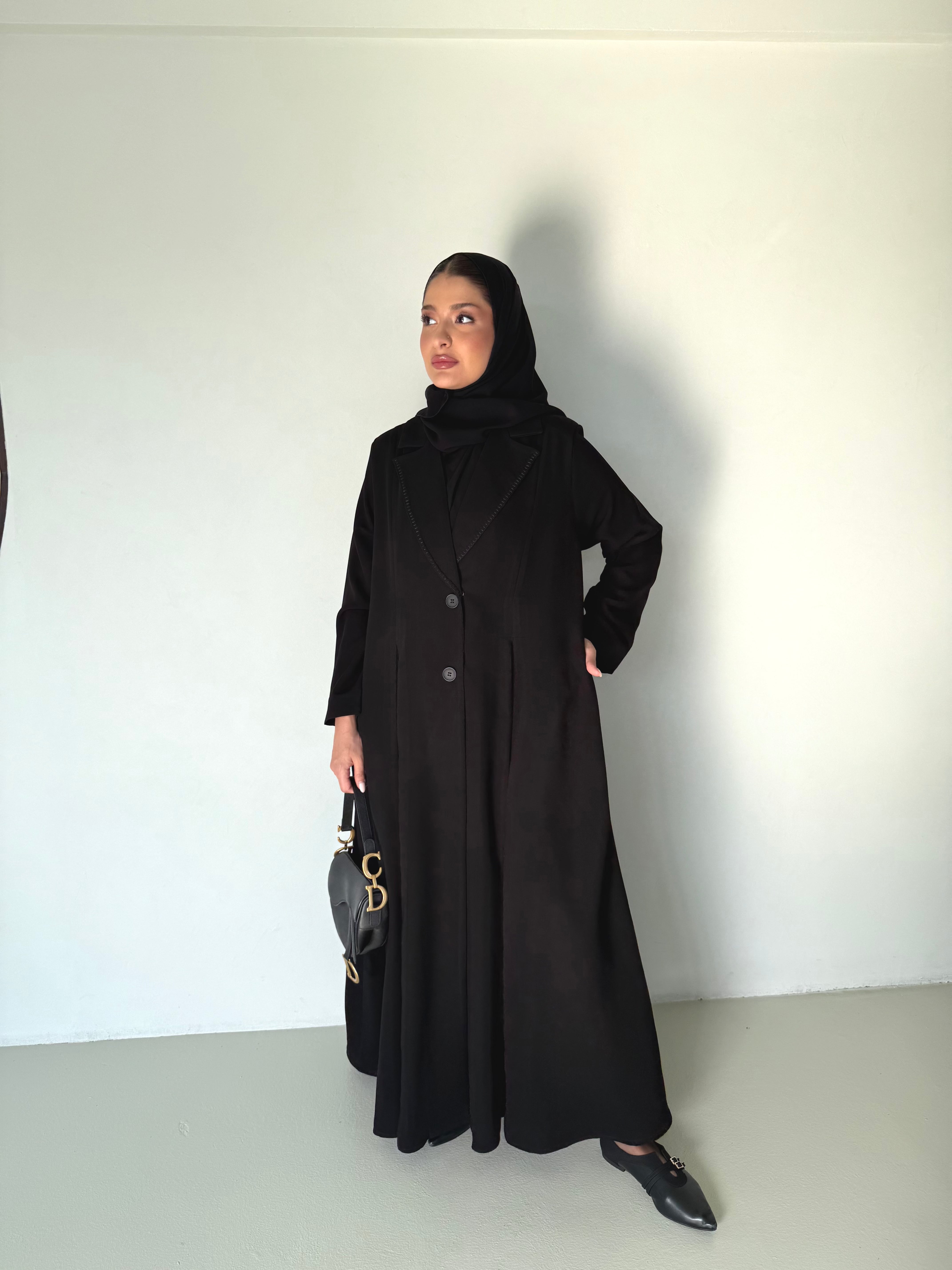 Woman wearing a black Modern Classic Crepe Abaya with tailored top and pleated skirt, holding a handbag.