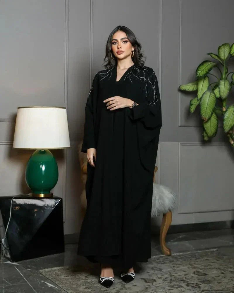Woman wearing black crepe bisht with silver and black hand embroidery on front and sleeves standing indoors next to a lamp and plant.