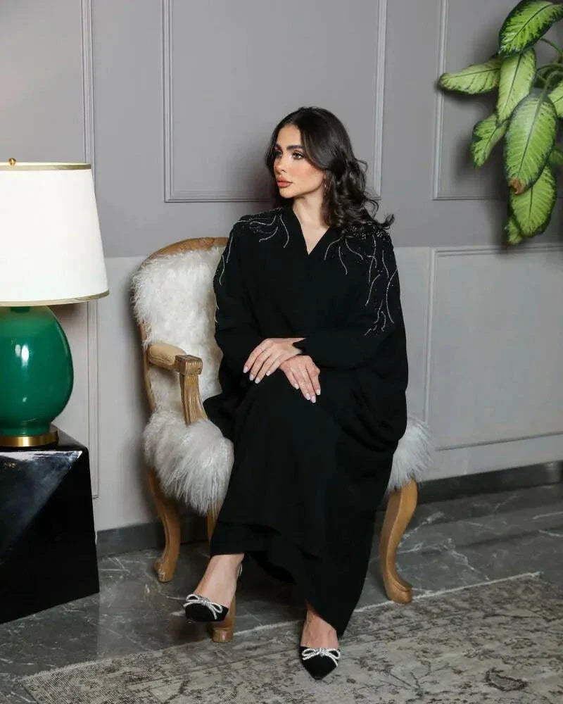 Woman wearing black crepe bisht with silver and black hand embroidery on front and sleeves standing indoors next to a lamp and plant.