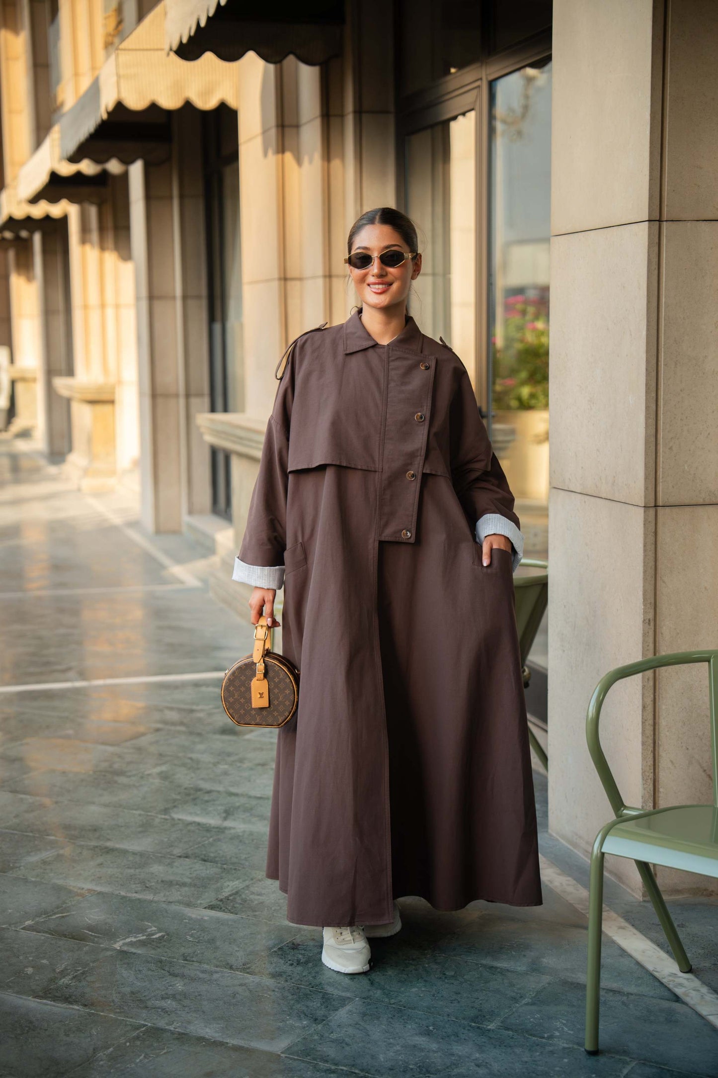 Woman wearing chocolate brown trench coat abaya with button-down front and layered detailing, styled with sunglasses and white sneakers outdoors
