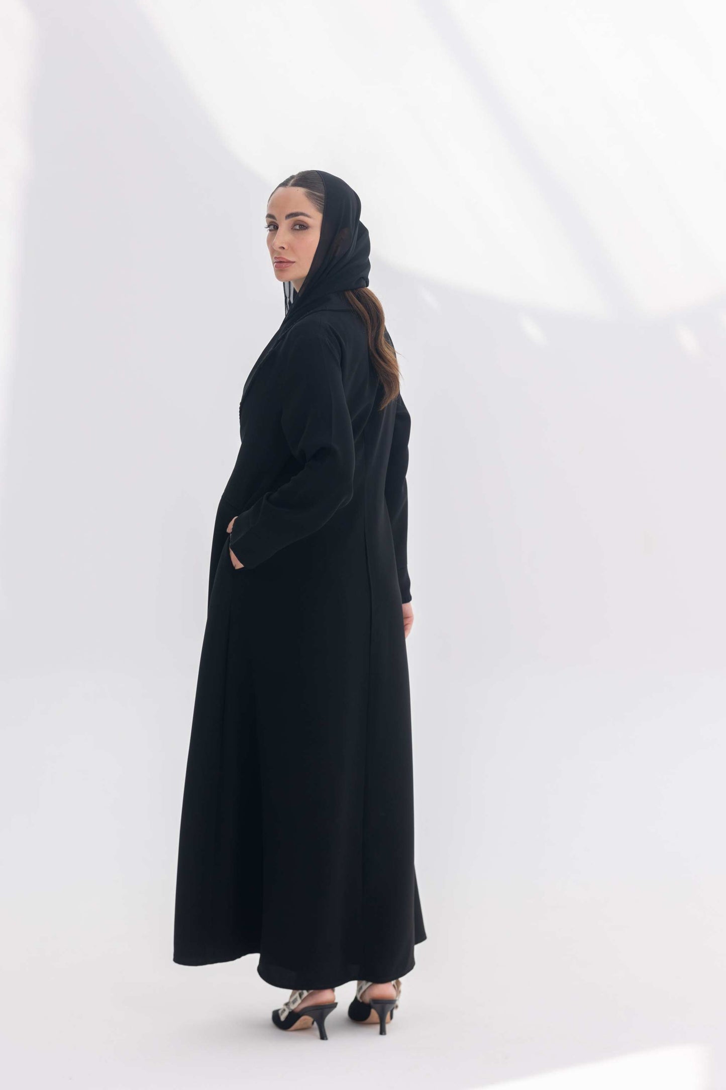 Woman wearing a sophisticated black crepe blazer abaya with tailored silhouette and black embroidery, paired with a matching black hijab and heels.