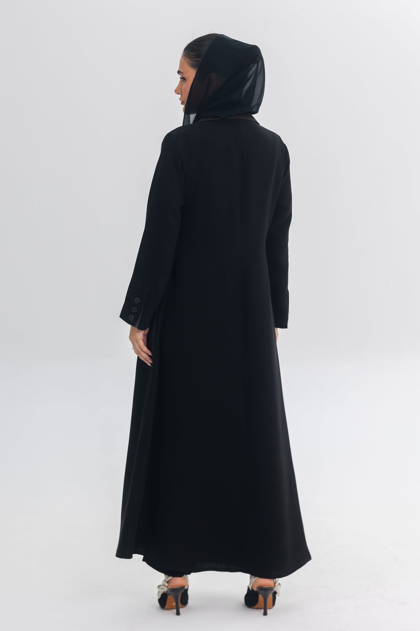 Back view of a woman wearing a sleek black crepe abaya with a hood, part of the Blue Stitch collection.