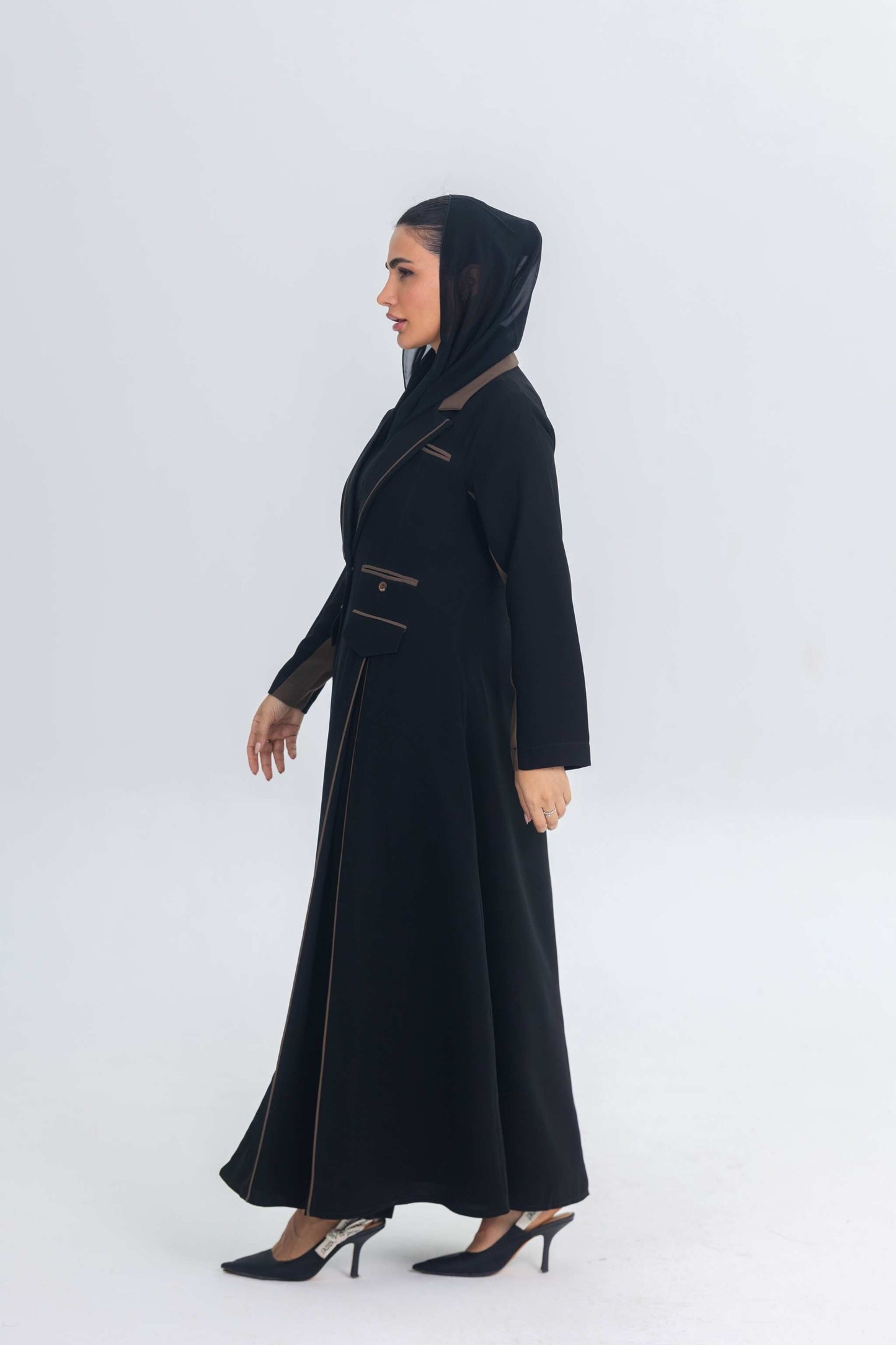 Woman wearing black crepe blazer abaya with chestnut brown trim and detailing, styled with matching hood and heels.