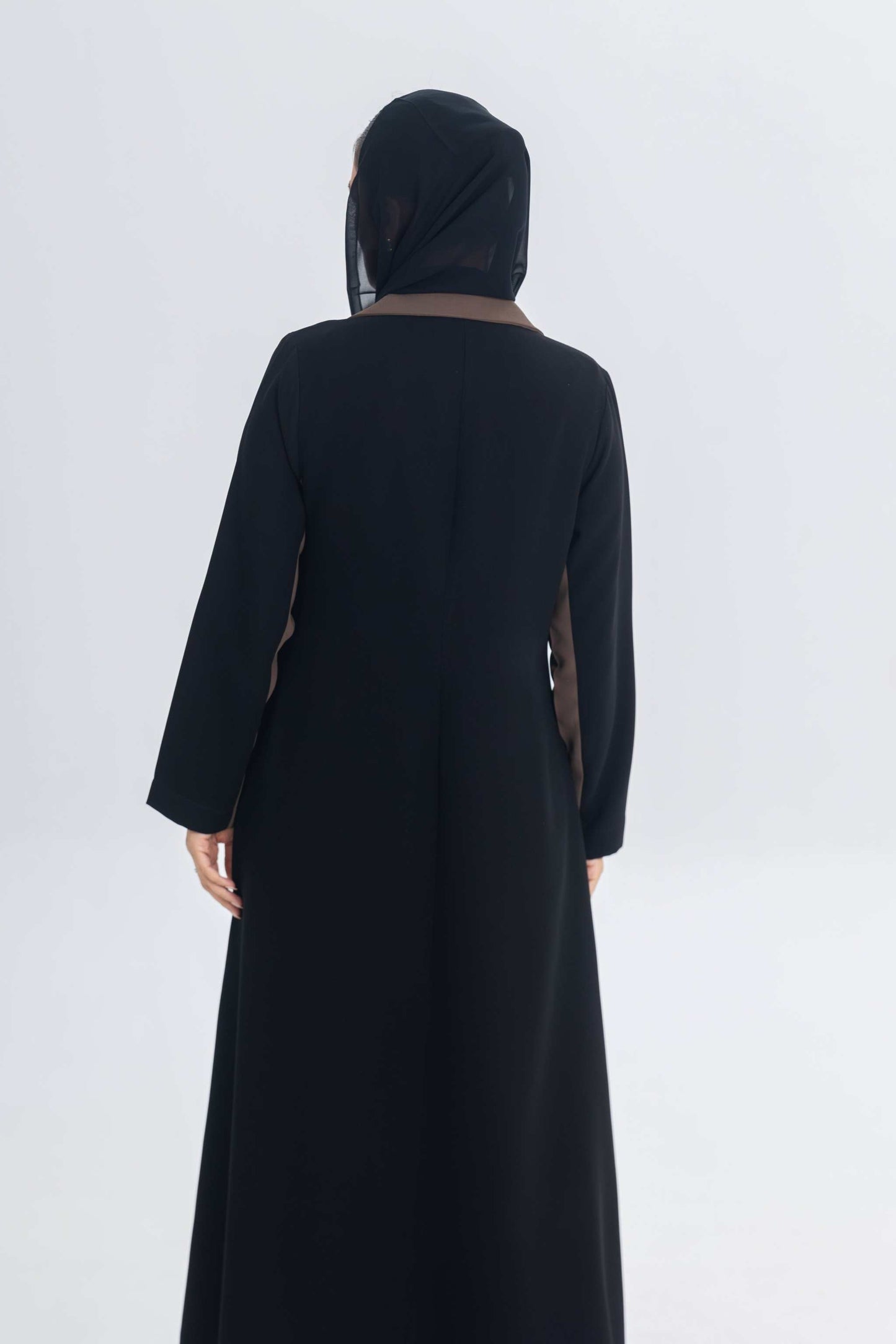 Back view of black crepe blazer abaya with chestnut brown trim worn with black headscarf on plain background