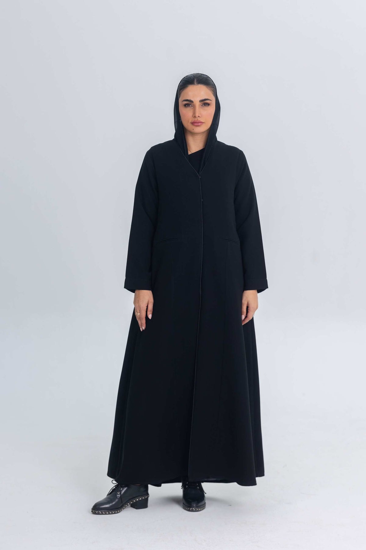 Woman wearing black crepe abaya with subtle satin detailing and hood, standing against plain background