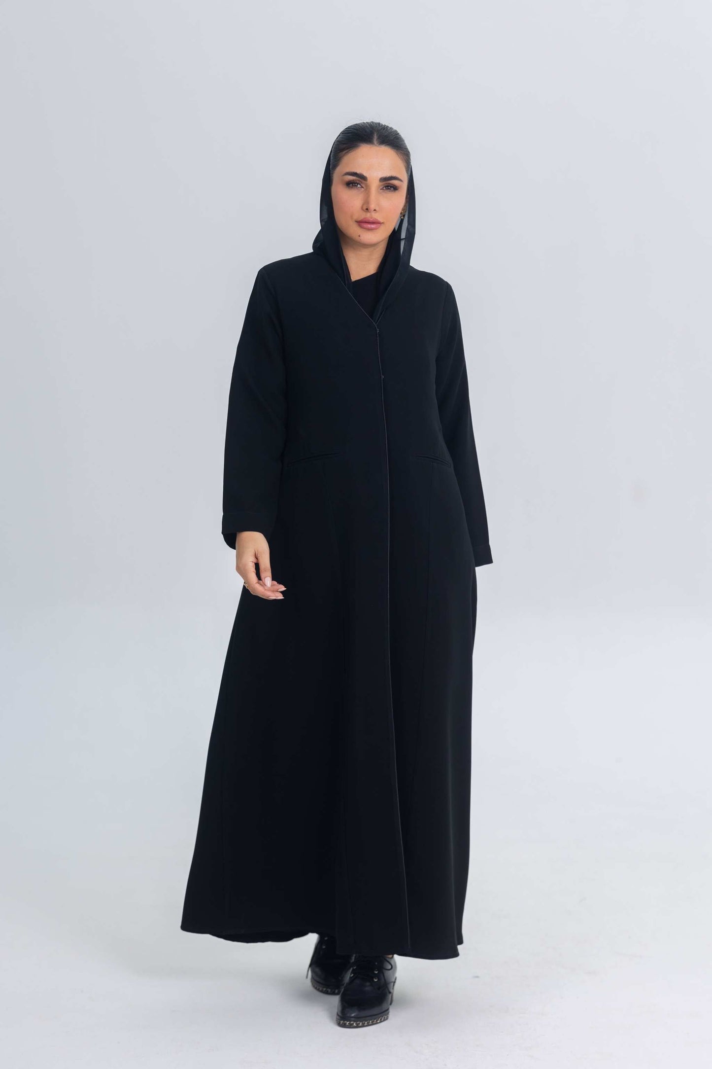 Woman wearing black crepe abaya with subtle satin detailing and hood, standing against plain background