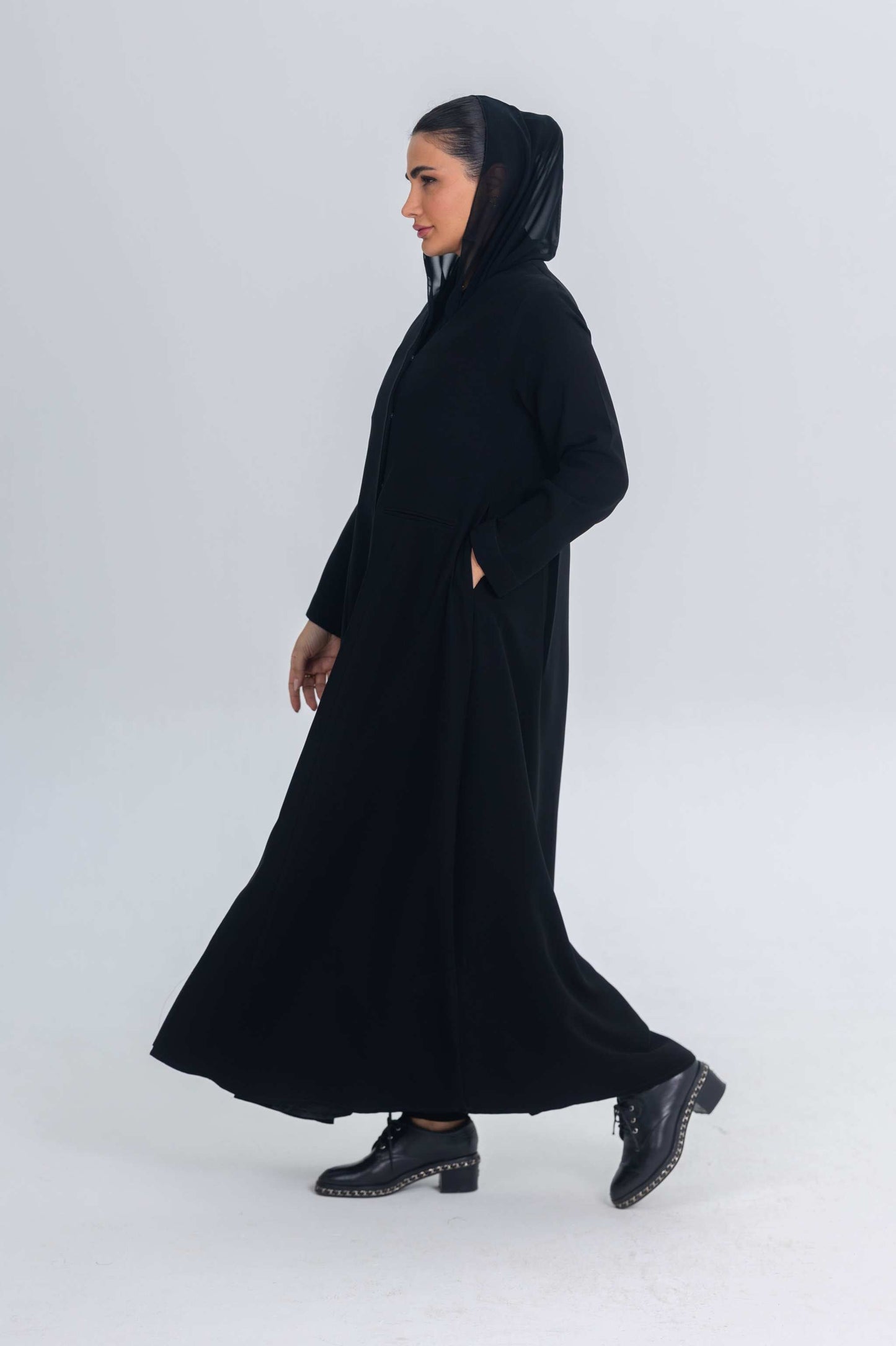 Woman wearing elegant black crepe abaya with satin detailing and hood walking against plain background