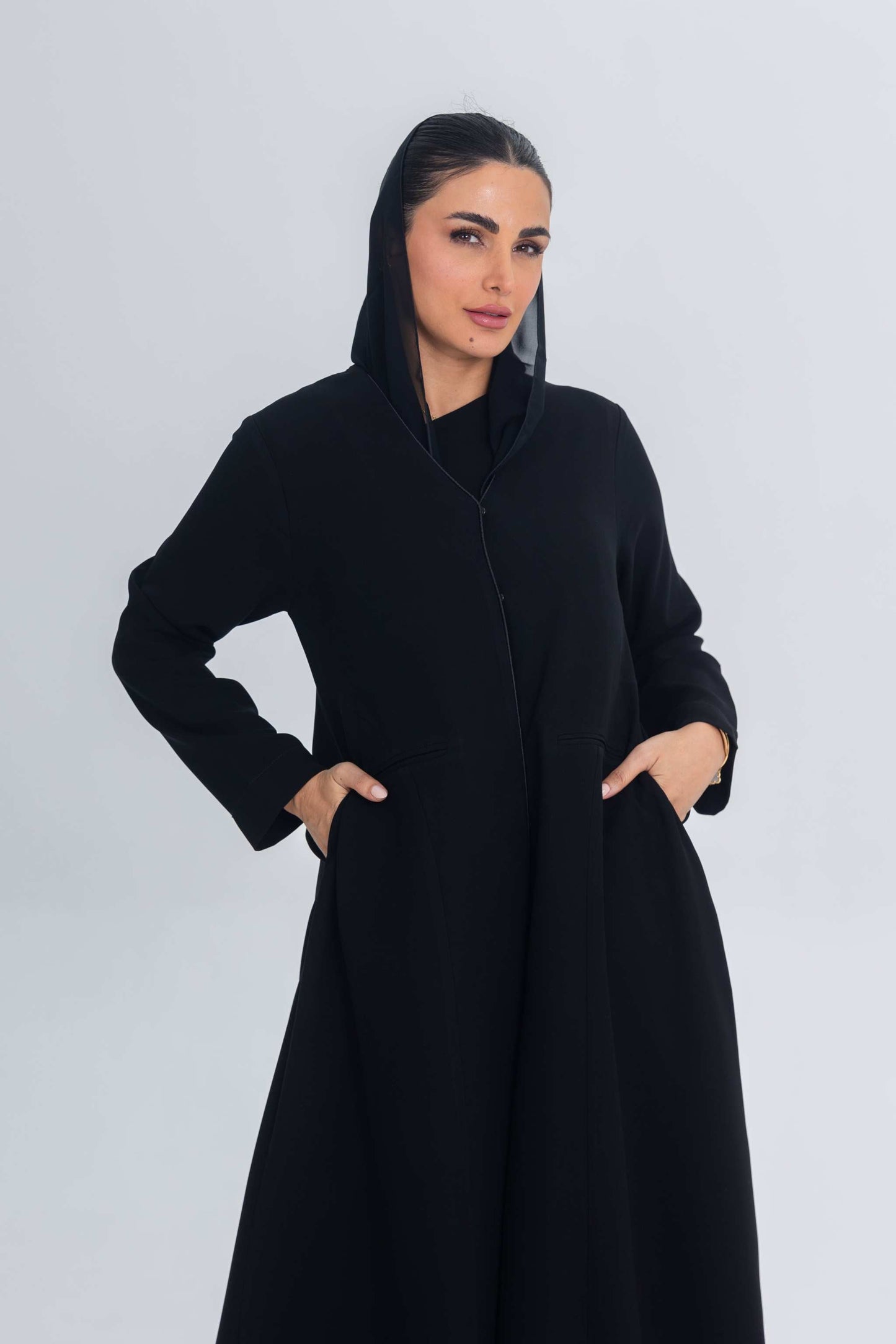 Woman wearing a black crepe abaya with subtle satin detailing and hood, hands in pockets, standing against a plain light background.