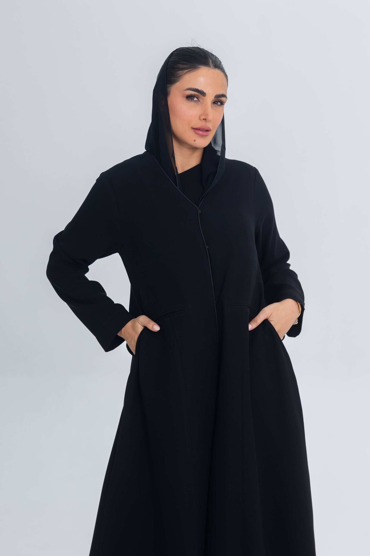 Woman wearing a black crepe abaya with subtle satin detailing and hood, hands in pockets, standing against a plain light background.