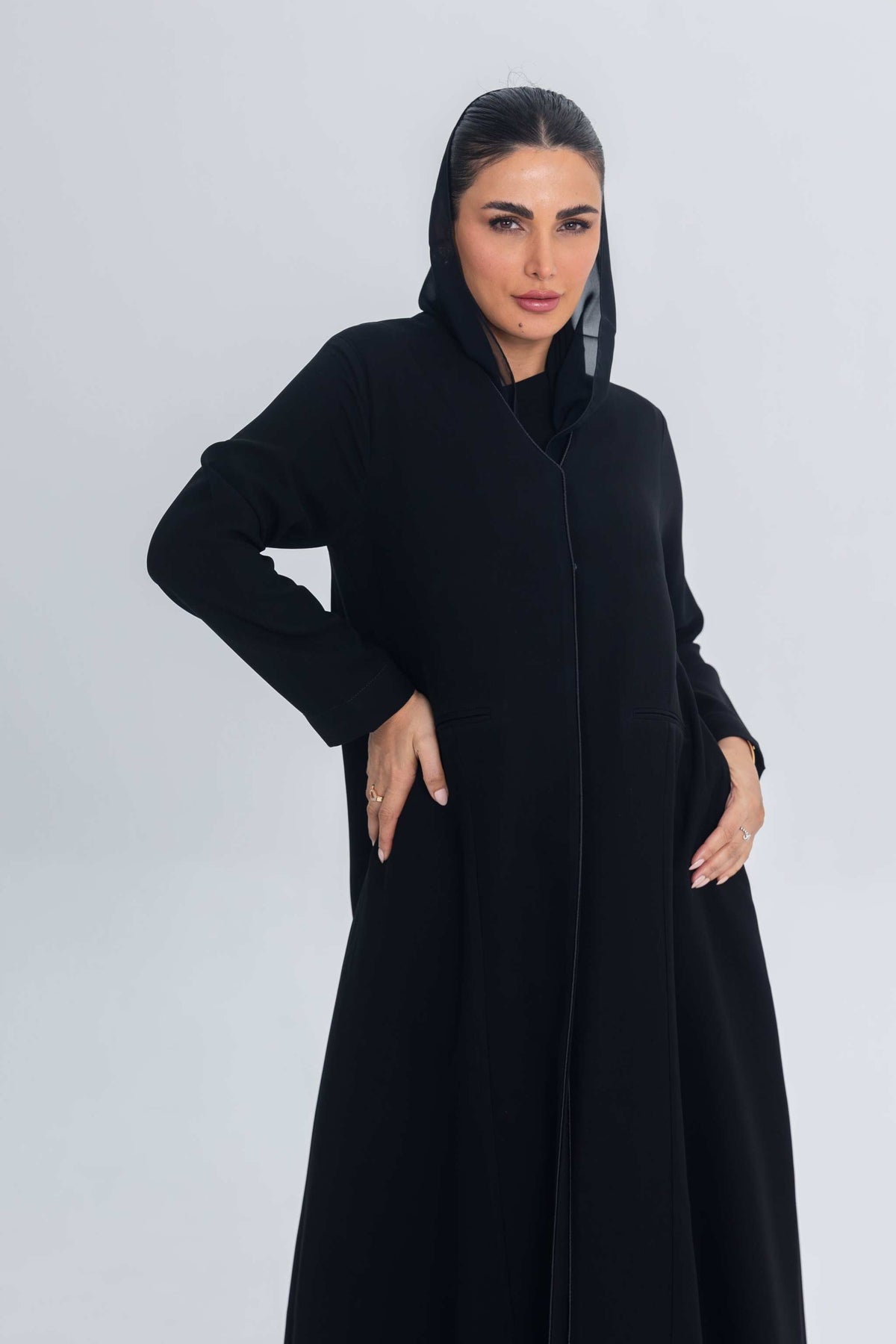 Woman wearing Noir Elan black crepe abaya with subtle satin detailing and hood against plain background