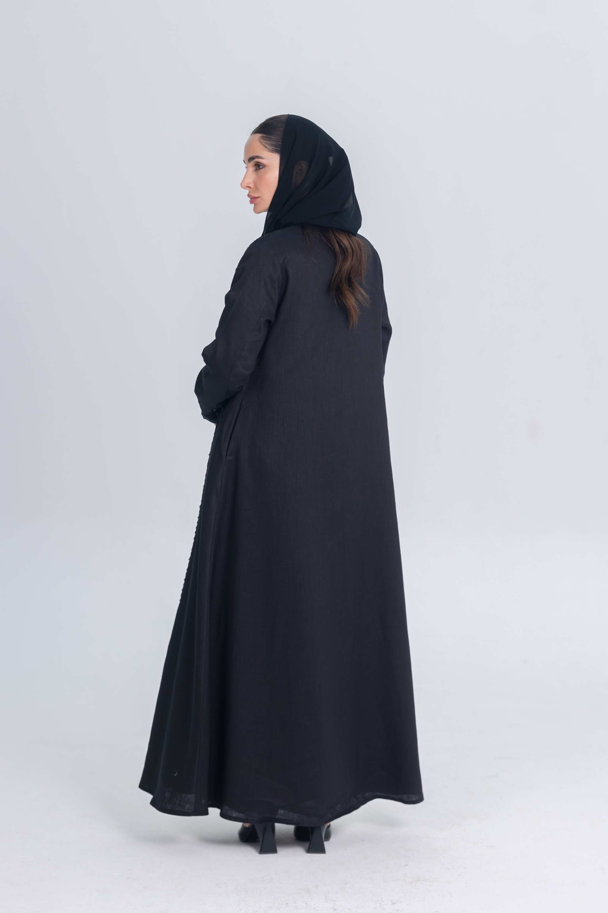 Woman wearing a black linen abaya with textured fabric detail and matching hijab, standing against a plain background.